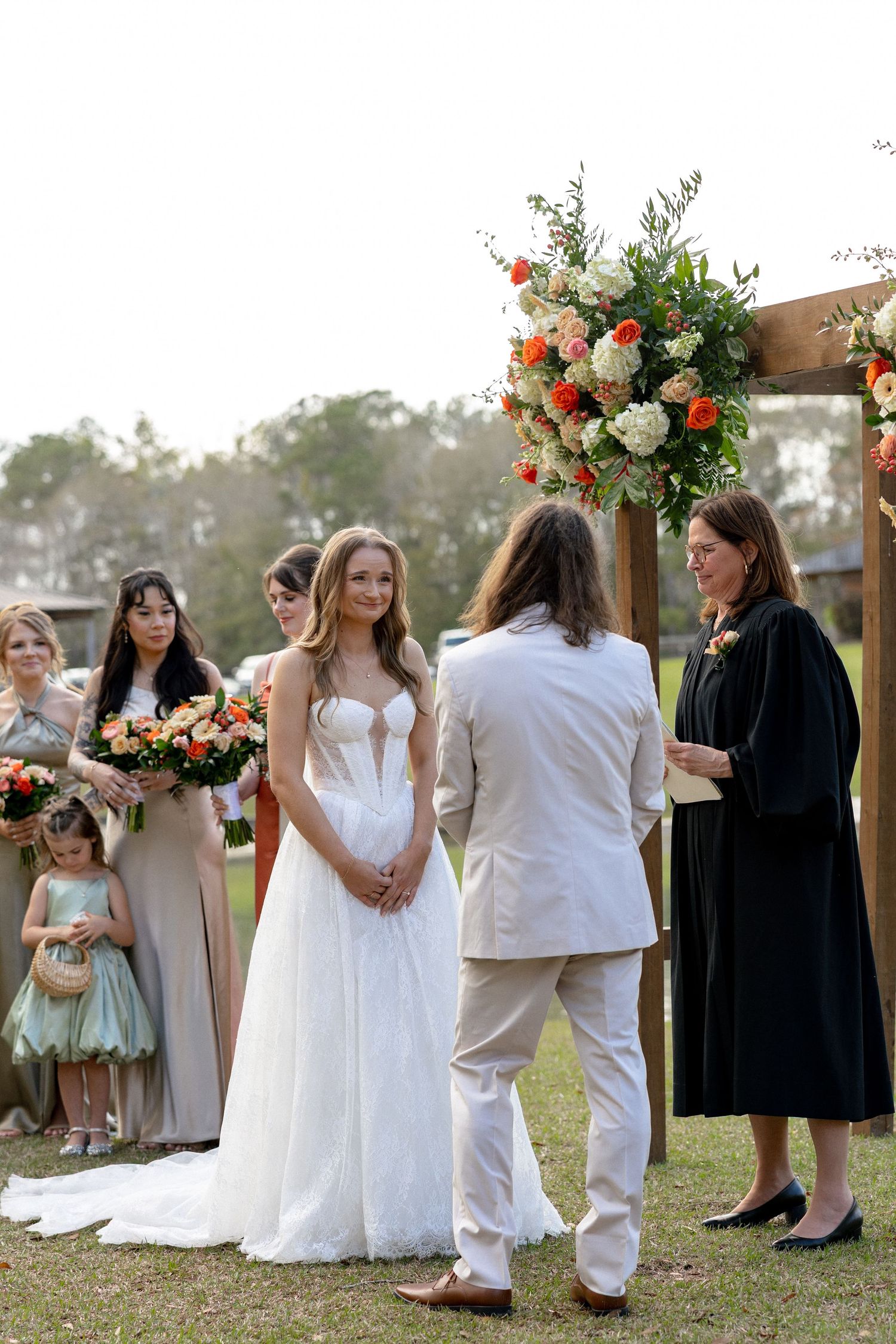 Bride and groom exchanging vows at outdoor wedding ceremony with bridesmaids and officiant under floral arch.