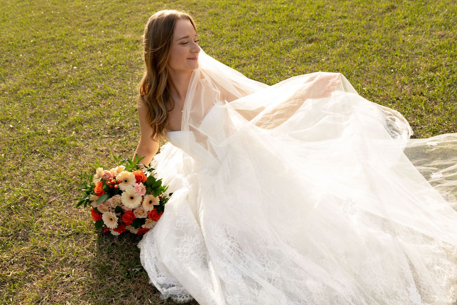 Bride in white lace wedding dress with veil holding bouquet on green lawn.