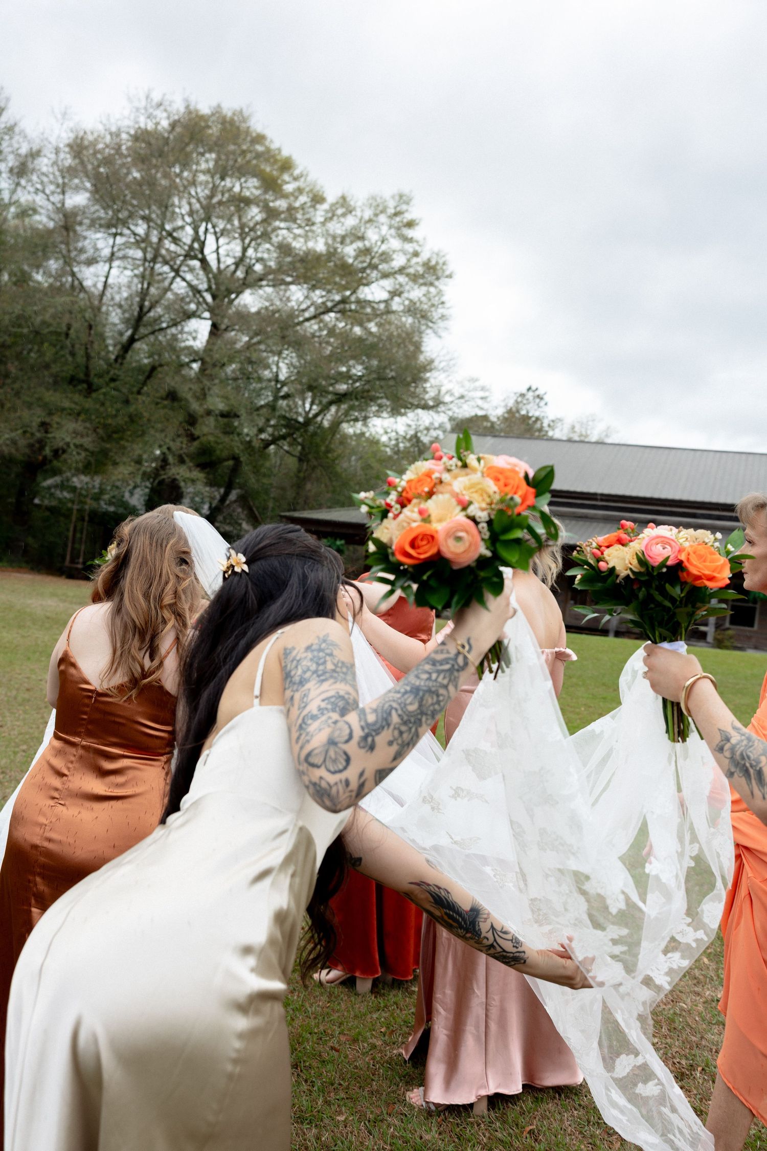 Tattooed bridesmaid in white dress adjusts bride's lace veil while bridal party gathers on green lawn.