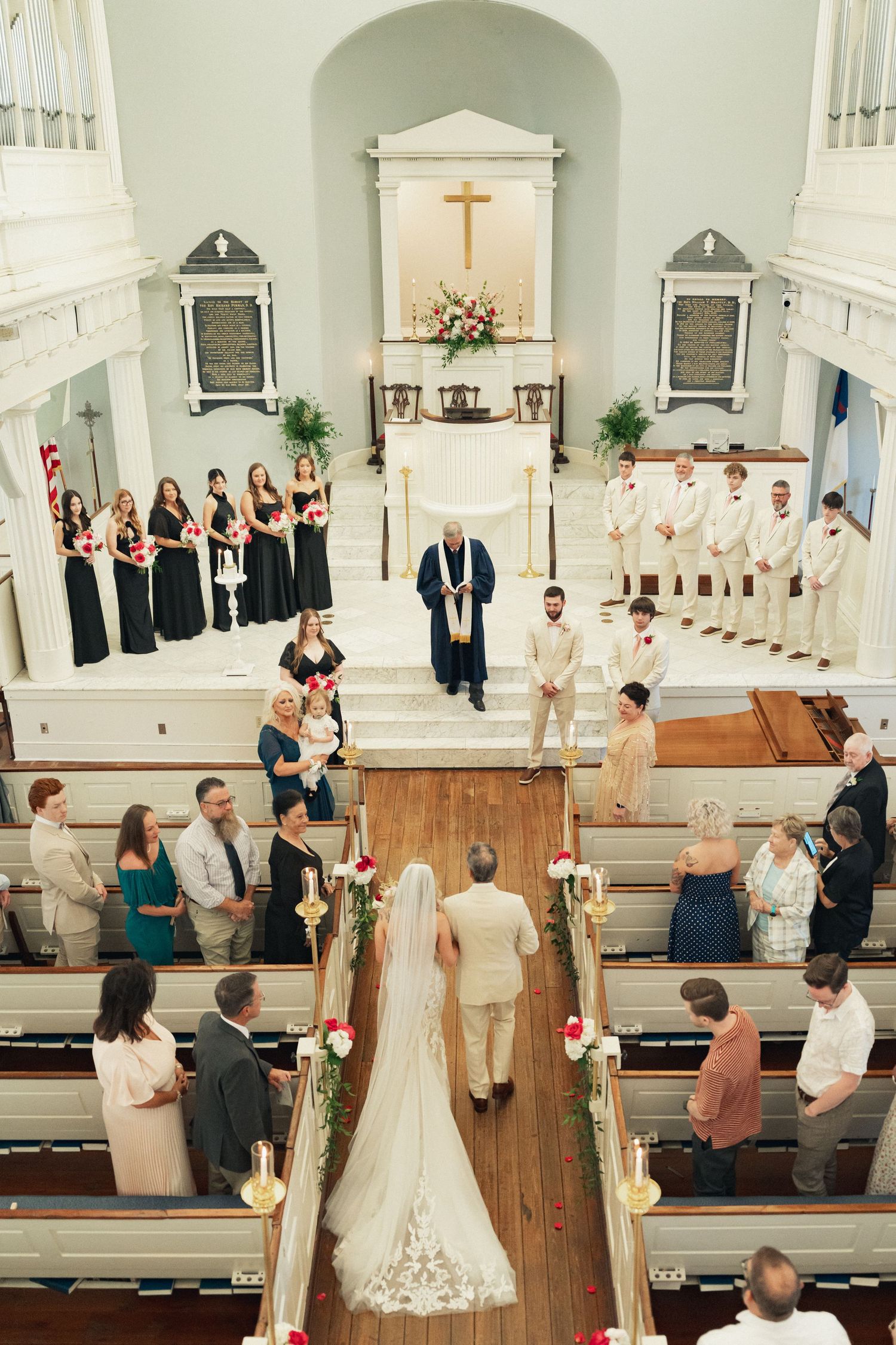 Aerial view of a church wedding ceremony with bride walking down the aisle, bridesmaids in black, groomsmen in white.