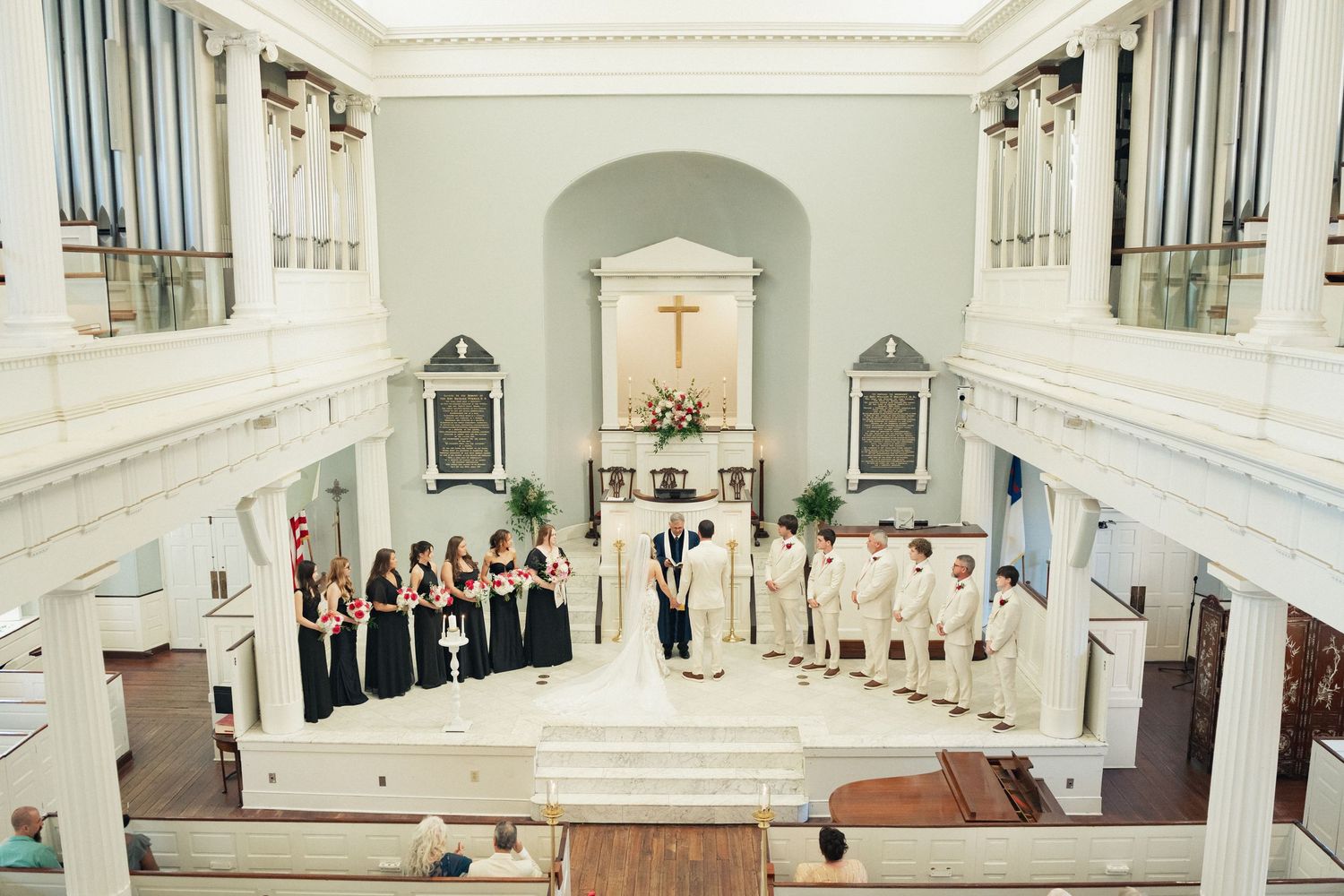 Wedding ceremony in elegant white church with bridesmaids in black gowns and groomsmen in white suits at the altar.