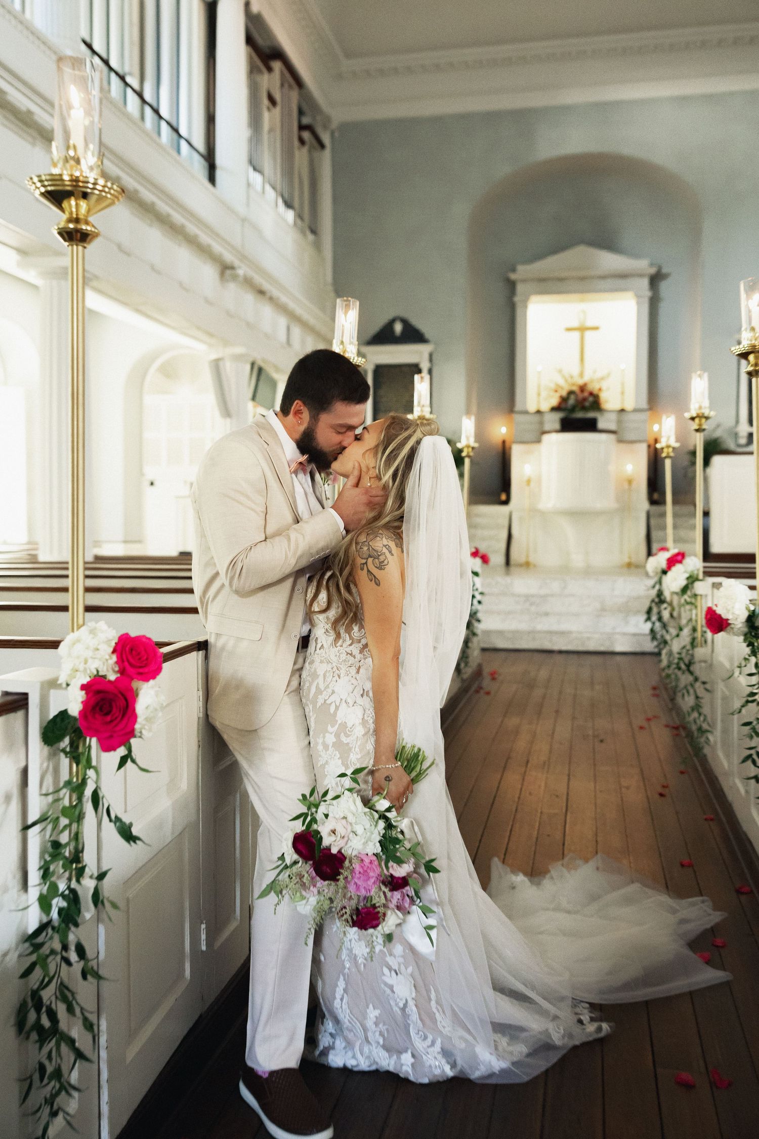 Bride and groom share first kiss in elegant church aisle adorned with red and white floral arrangements.