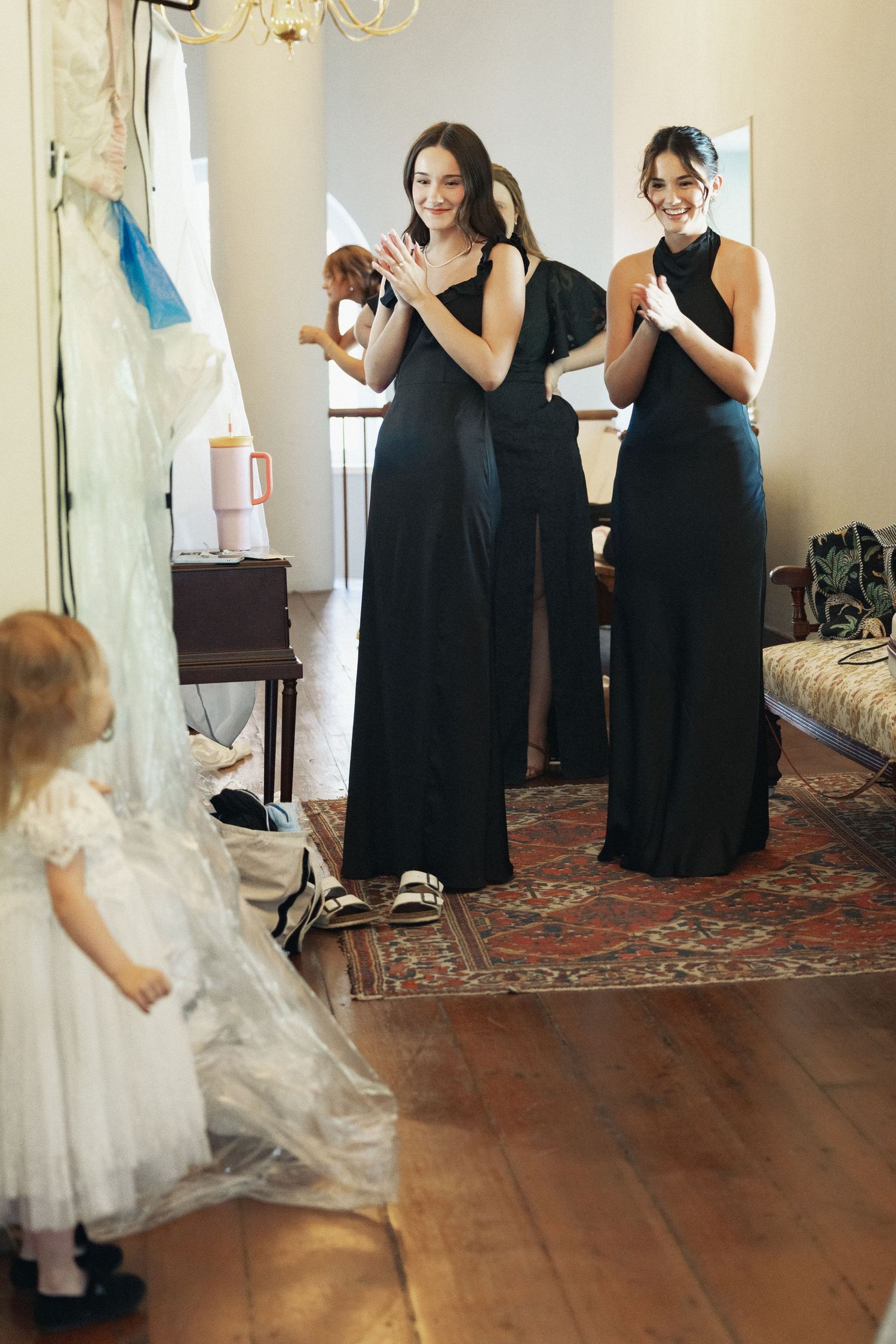 Two smiling bridesmaids in black gowns admire a flower girl in white dress during wedding prep.