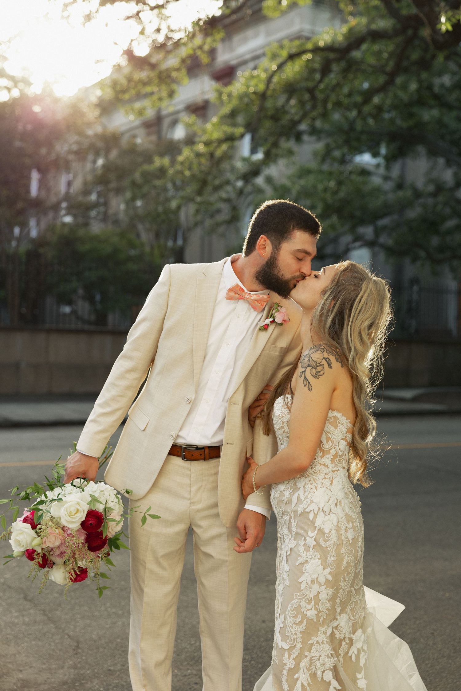 Bride and groom share a romantic kiss on a sunlit street, groom in beige suit holding a red and white bouquet.