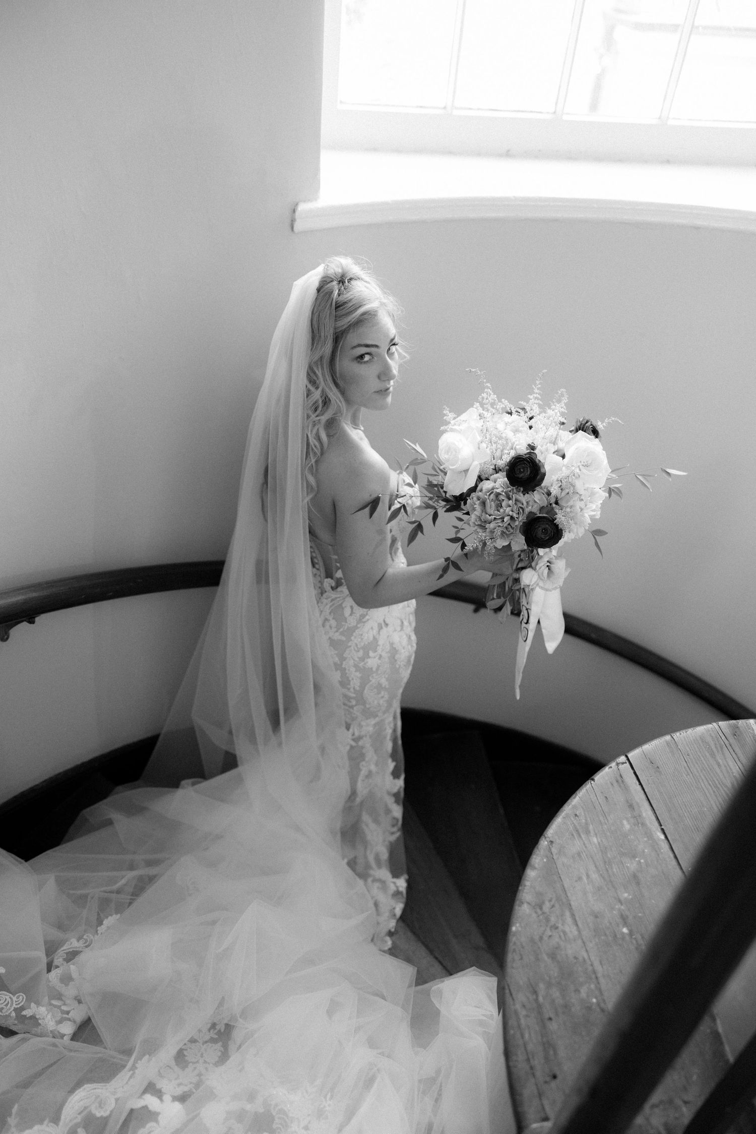 Black and white photo of bride in lace gown and veil on spiral staircase holding bouquet.