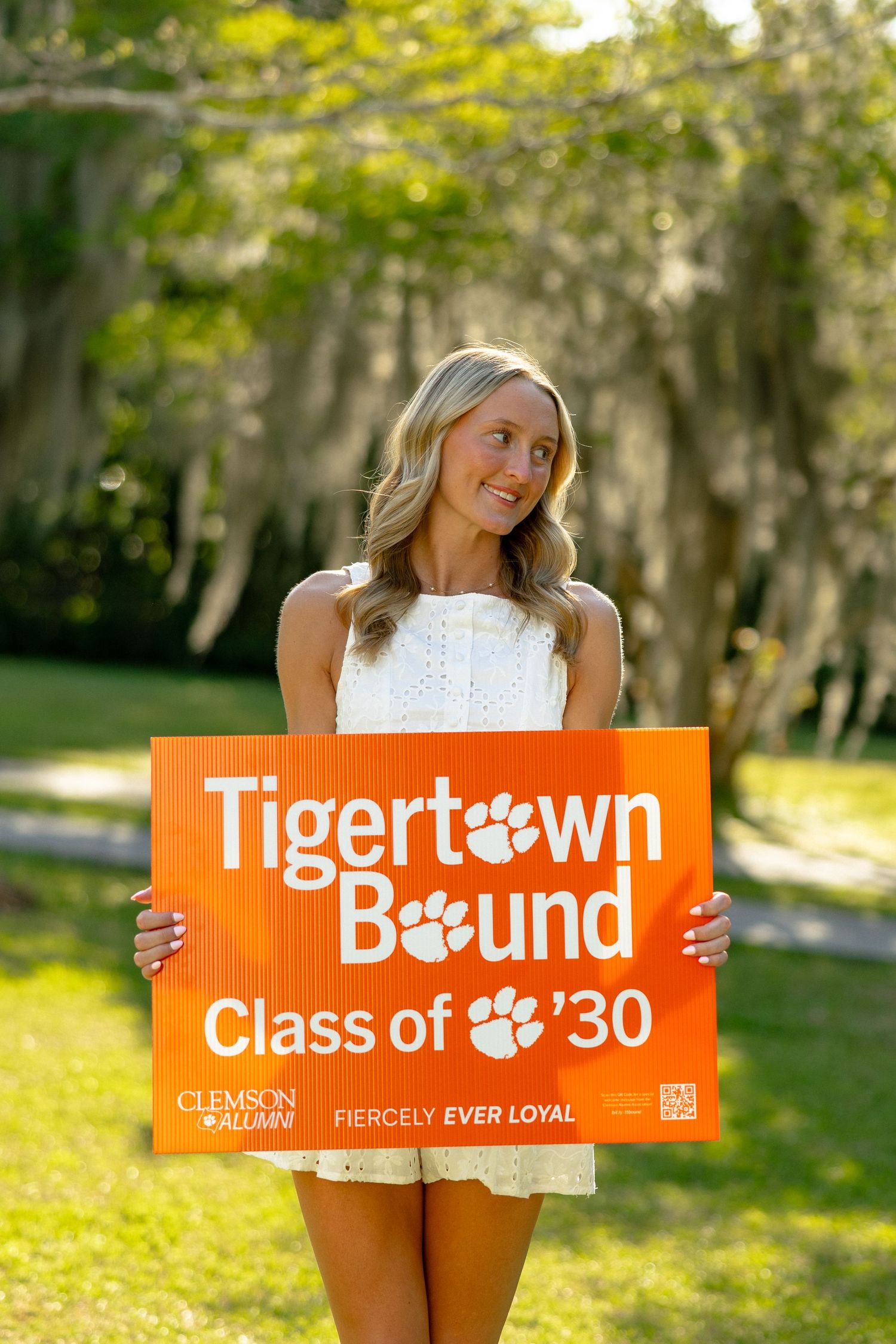 Smiling blonde woman holding a Clemson Tigertown Bound Class of 2030 orange sign in a sunny park.