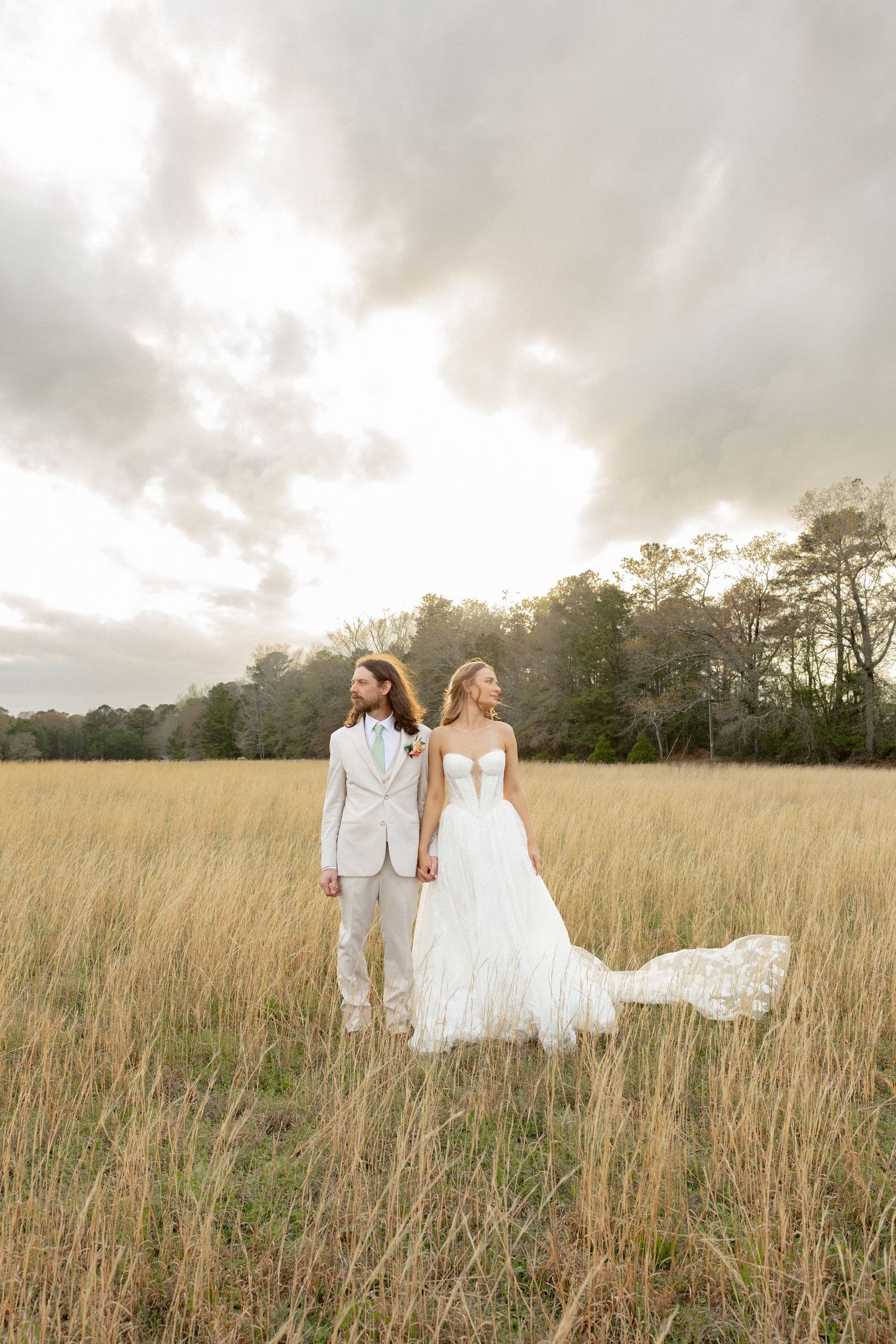 Bride and groom hold hands in golden meadow, bride's lace gown train flowing behind her.