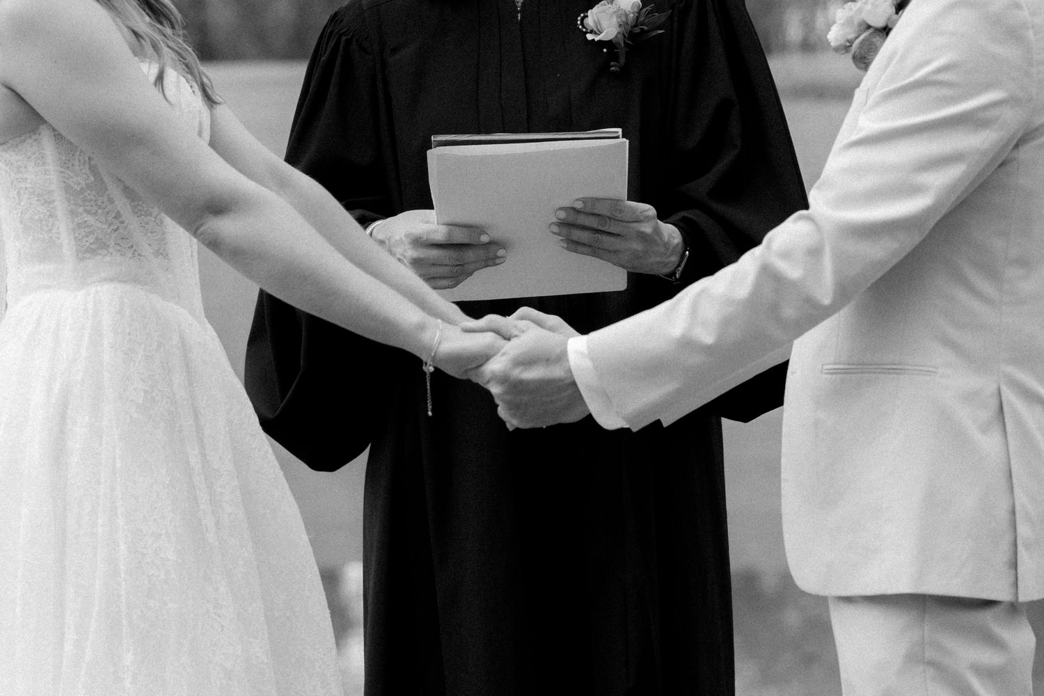 Black and white close-up of bride and groom holding hands during wedding ceremony with officiant.