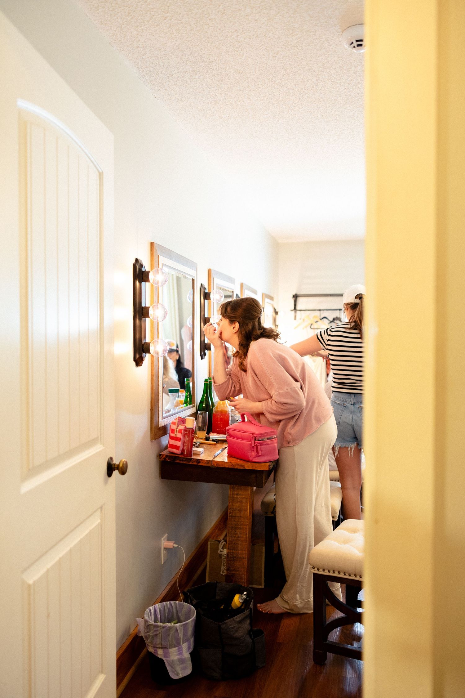 Two women getting ready at a vanity mirror with Hollywood lights during bridal preparations.