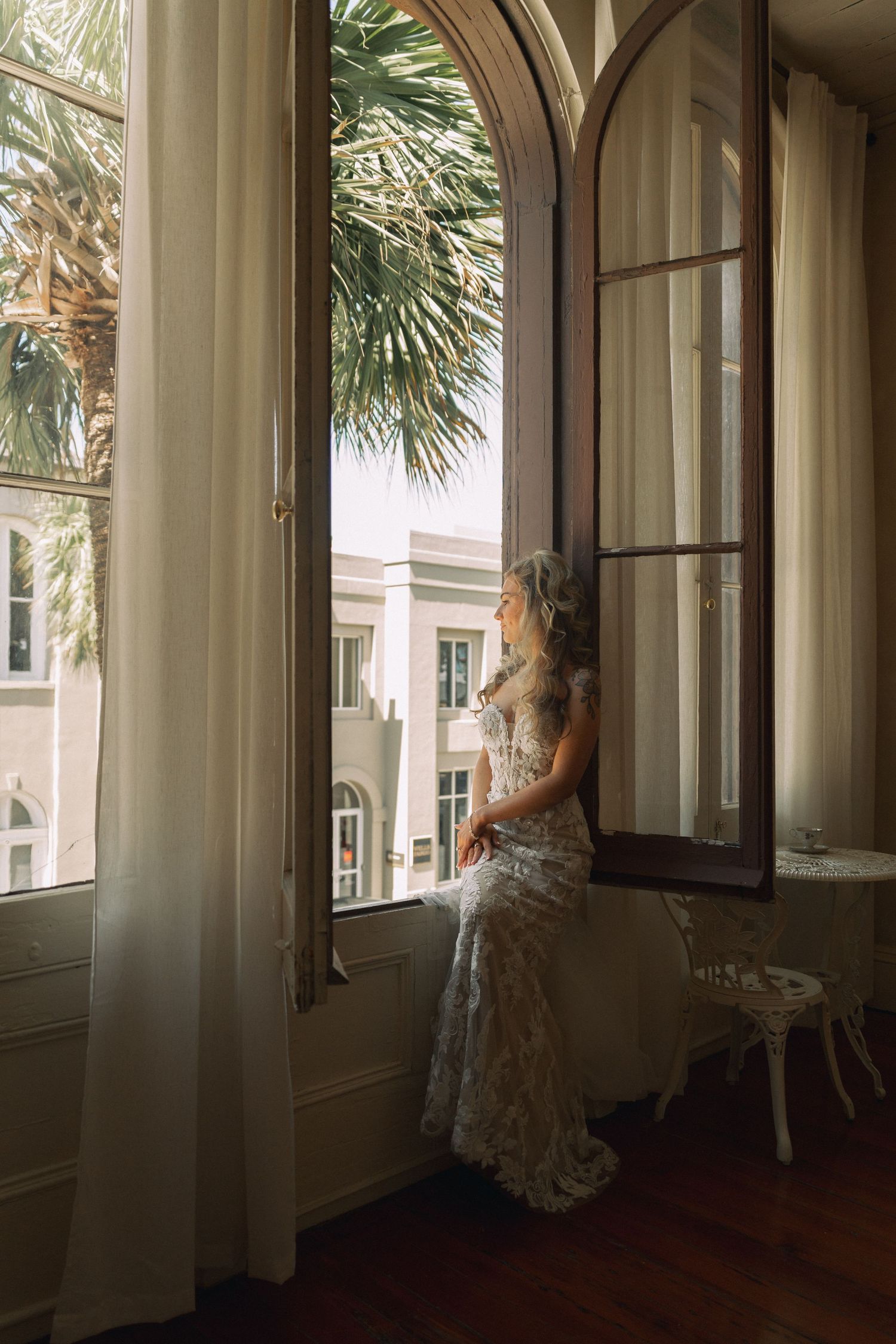Bride in lace wedding dress sitting on window sill gazing outside at palm trees in elegant historic venue.
