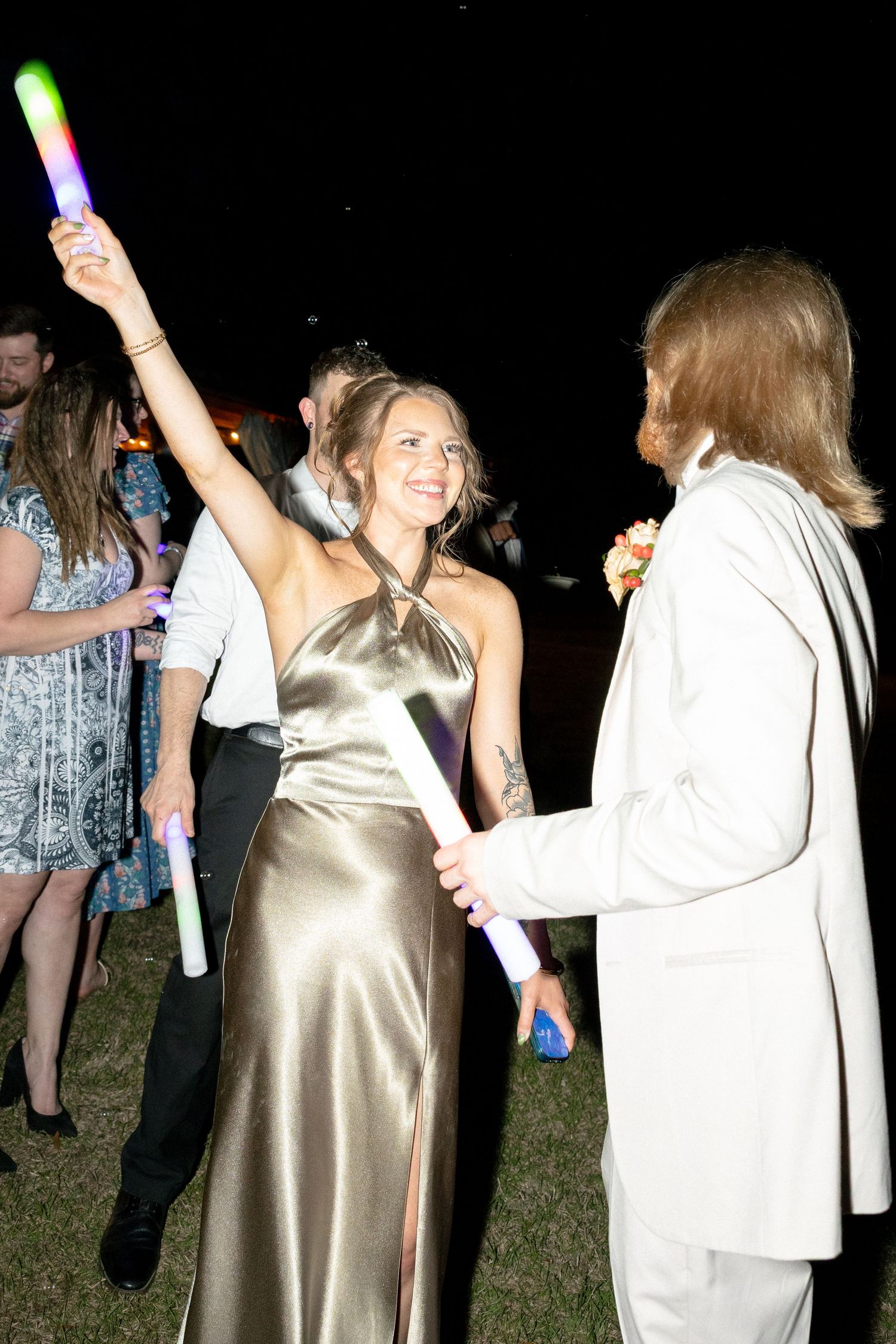 Young woman in gold satin gown raises glow stick at prom night dance, facing partner in white suit outdoors.