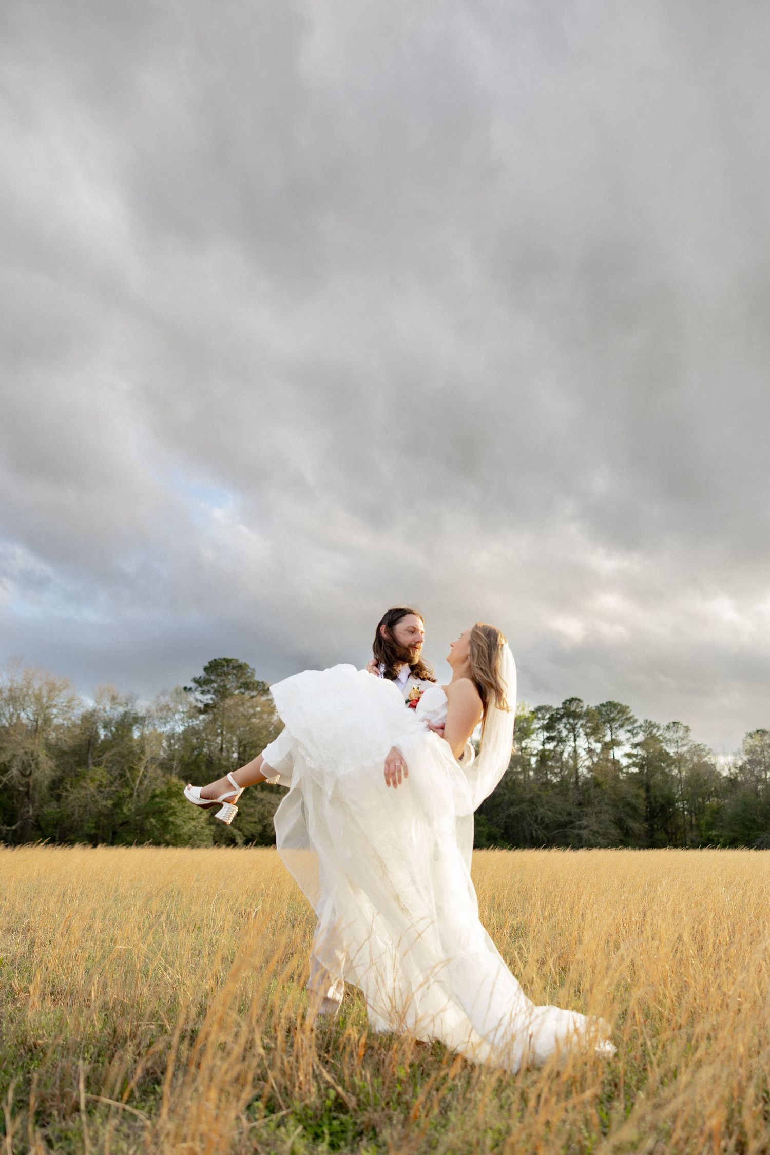 Groom carries bride in white gown through golden field under dramatic cloudy sky.