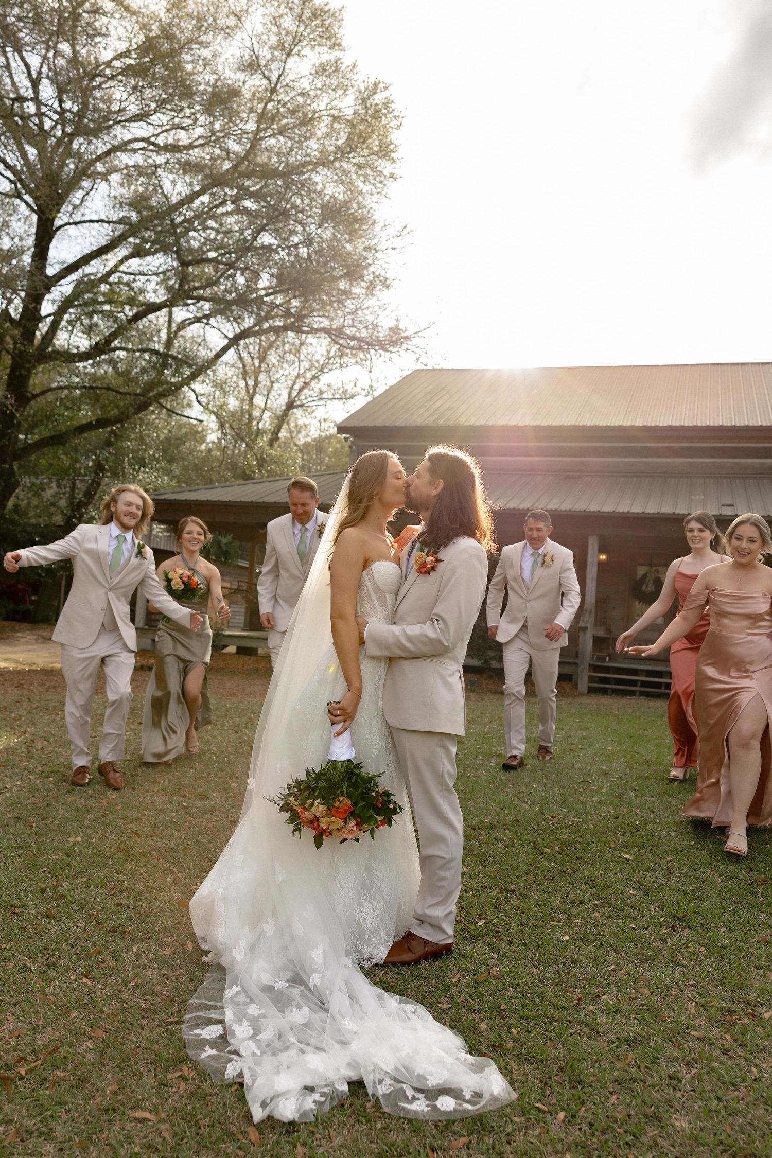 Bride and groom kiss while wedding party walks away in golden afternoon sunlight.