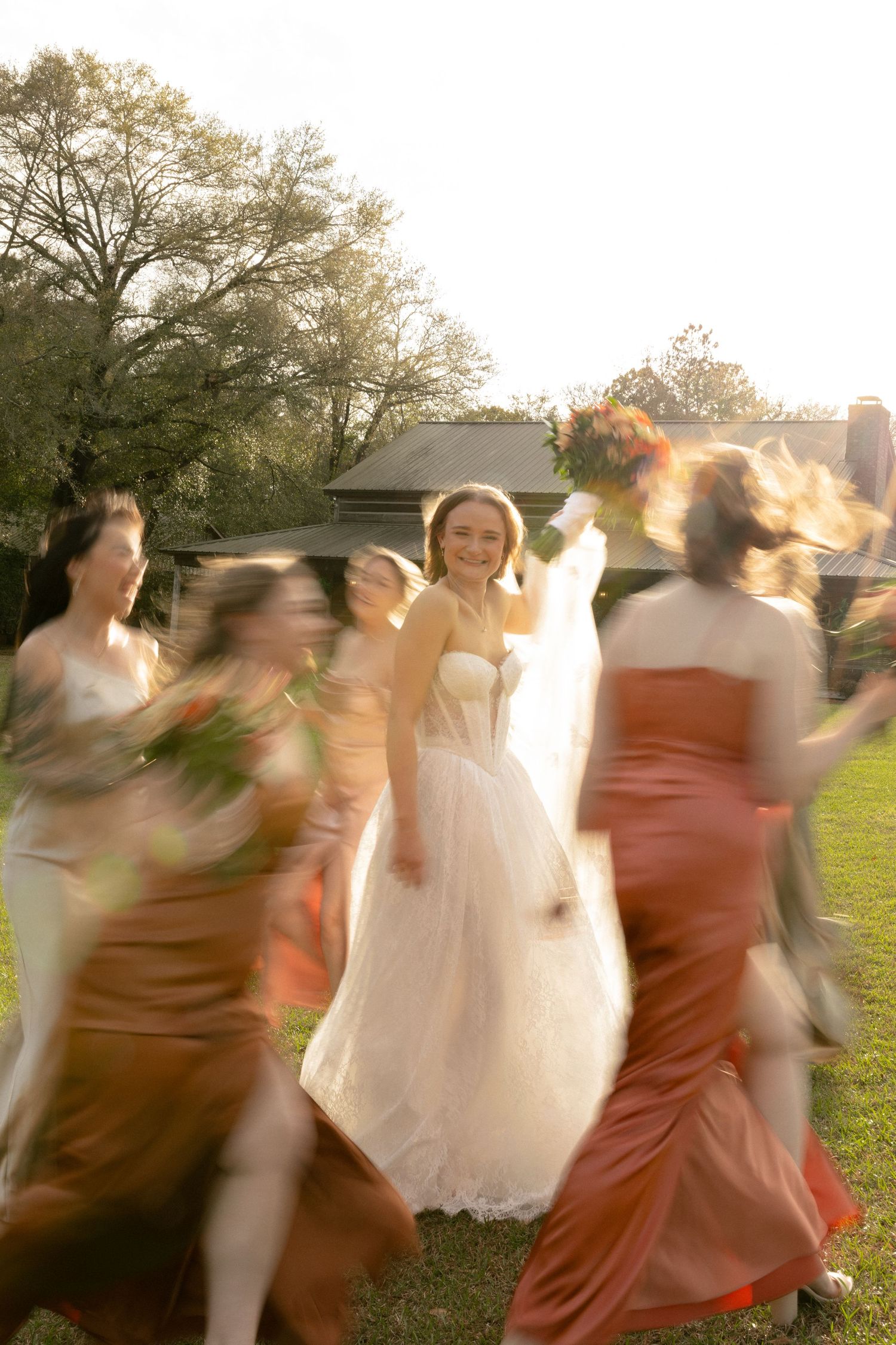 Bride smiling in white strapless gown surrounded by motion-blurred bridesmaids in rust and gold dresses.