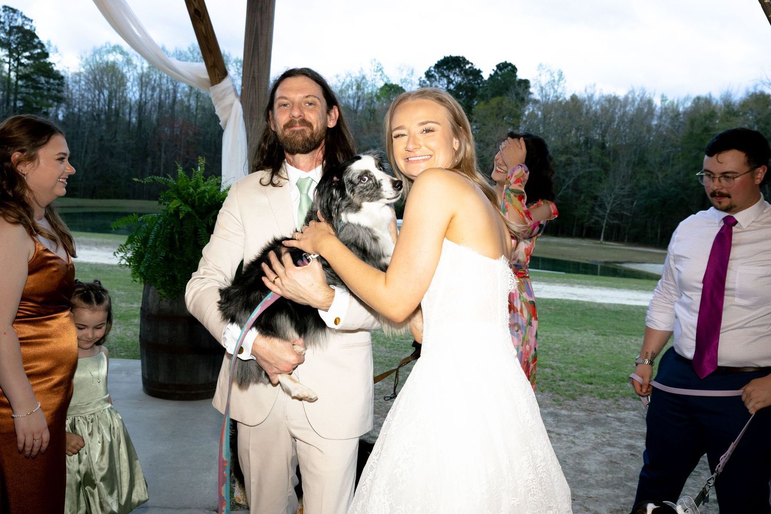 Smiling bride and groom pose with their dog at an outdoor wedding reception surrounded by guests.