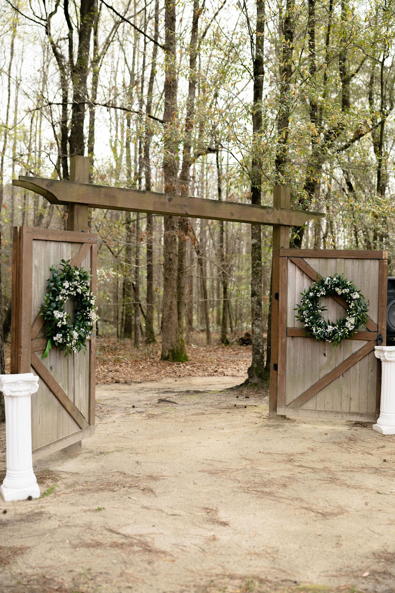 Rustic wooden wedding entrance gate adorned with two green wreaths opening to a wooded forest path.