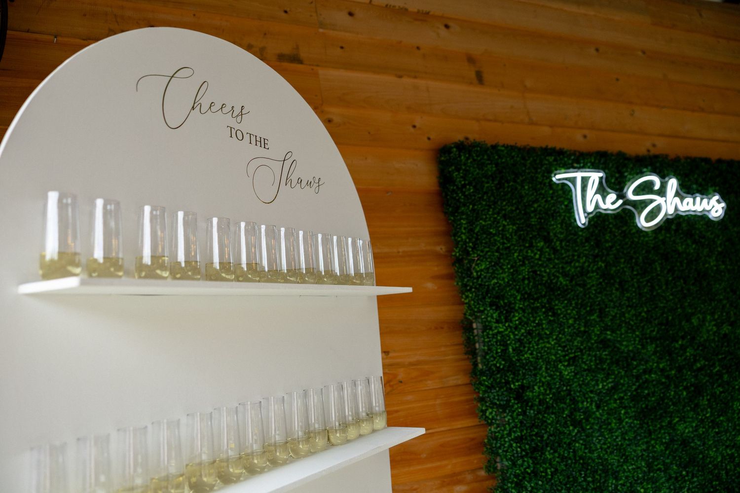 Champagne wall display reading Cheers to the Shaws beside a neon sign on a moss wall backdrop.