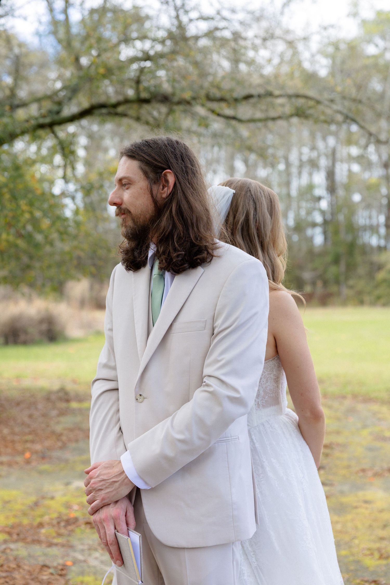 Bearded groom in beige suit with green tie stands back to back with bride holding vows card outdoors.