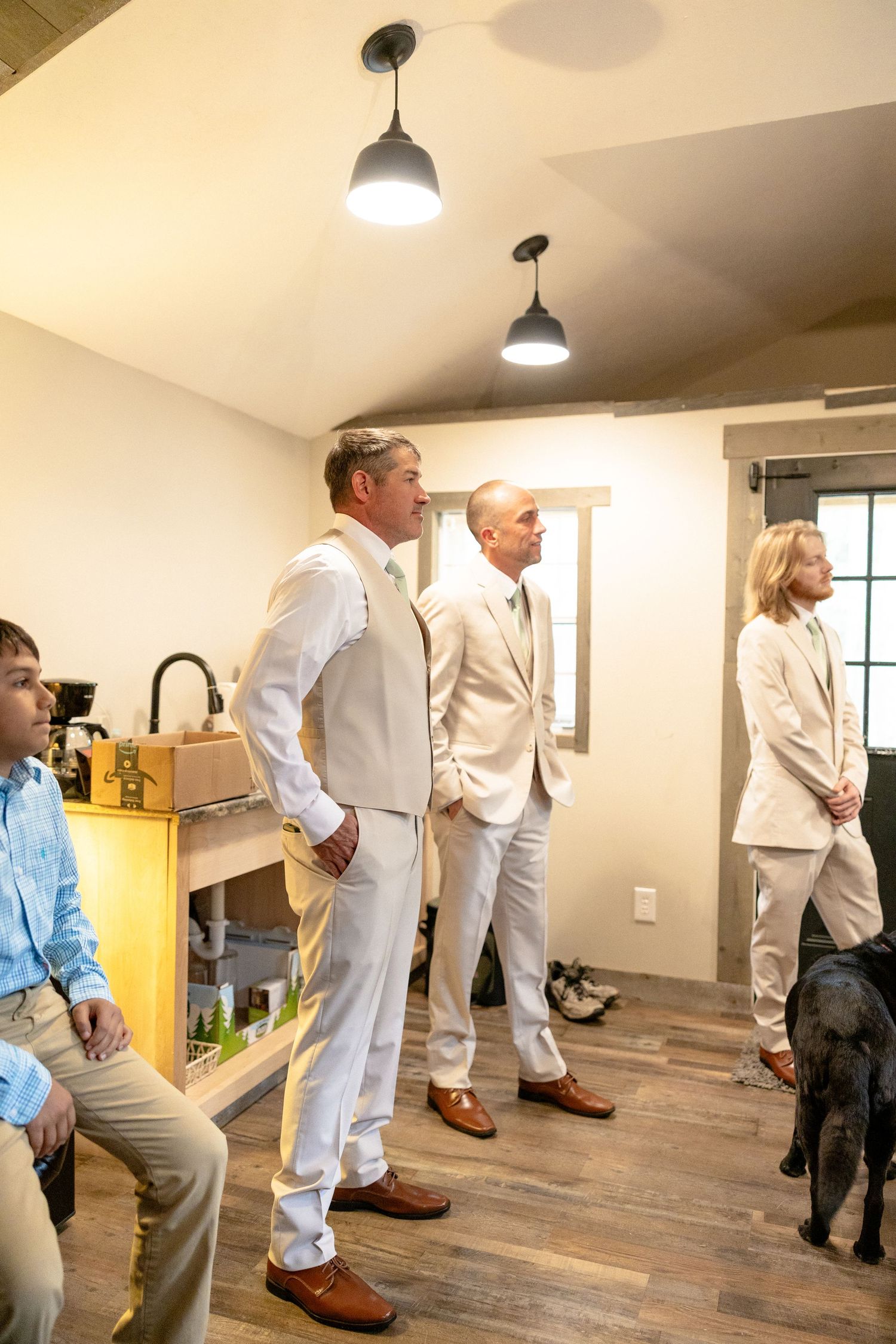 Groomsmen in beige suits with brown shoes gather in a rustic room before a wedding ceremony.