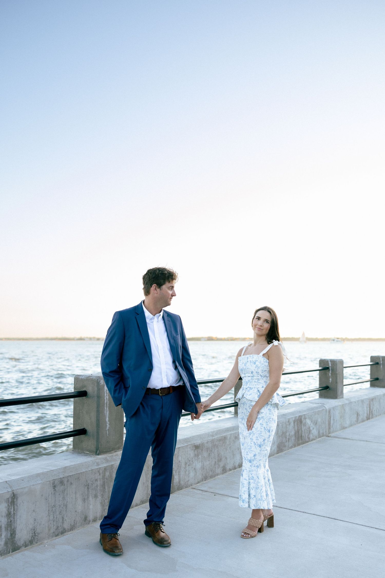 Couple holding hands on waterfront promenade at sunset, man in blue suit, woman in floral dress.