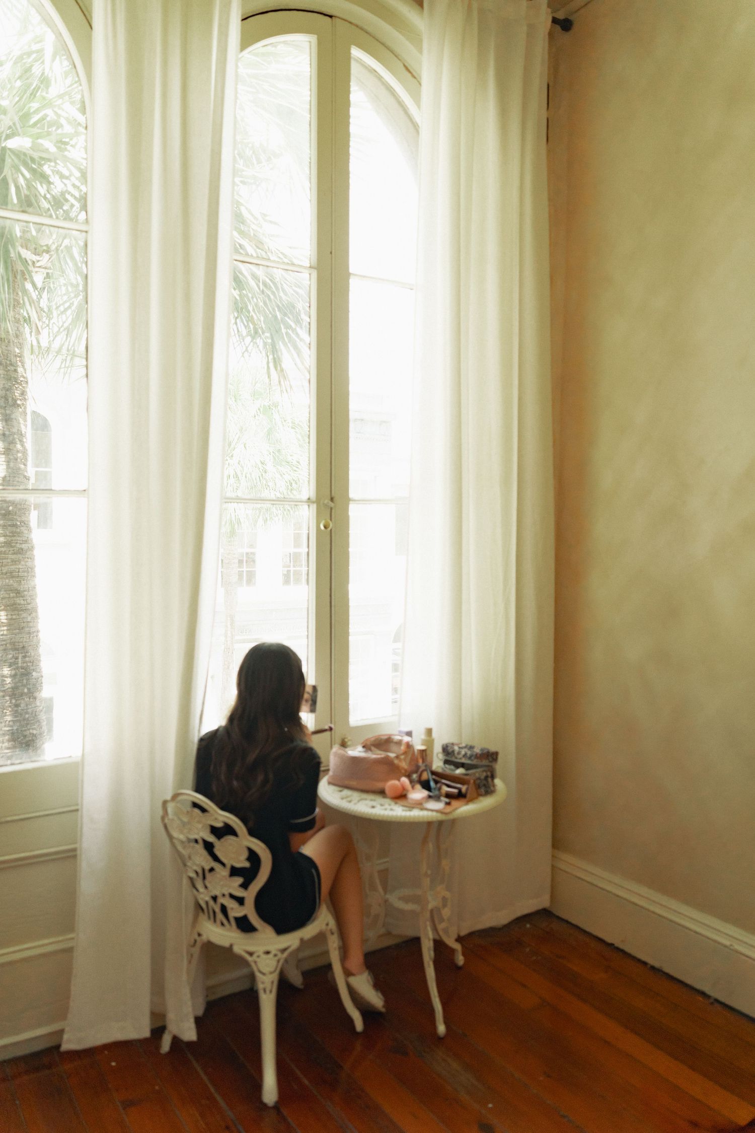 Woman in black dress sits at white ornate vanity table by tall arched window with white curtains on hardwood floor.