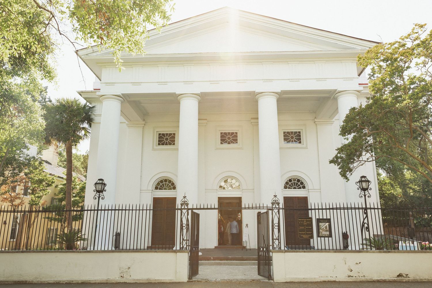White Greek Revival building with grand columns, iron fence, and sunlit trees in background.