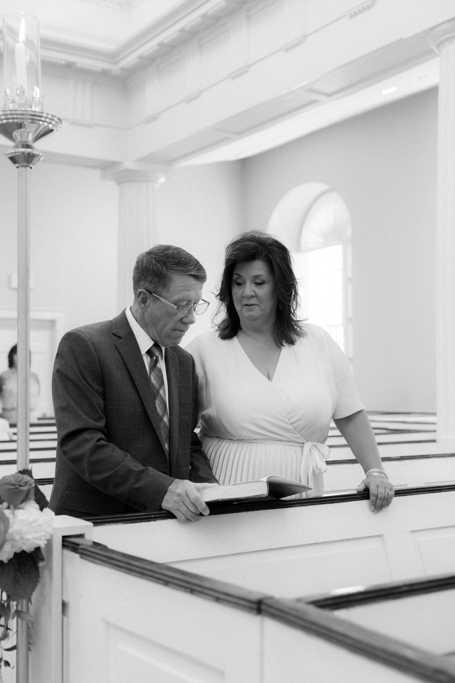 Black and white photo of a couple reviewing the wedding program together in a white church pew.