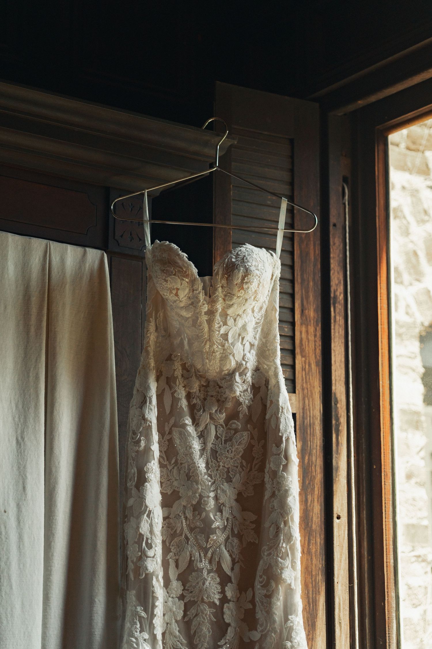 Lace wedding dress on a wire hanger displayed in a rustic wooden doorway with soft natural light.