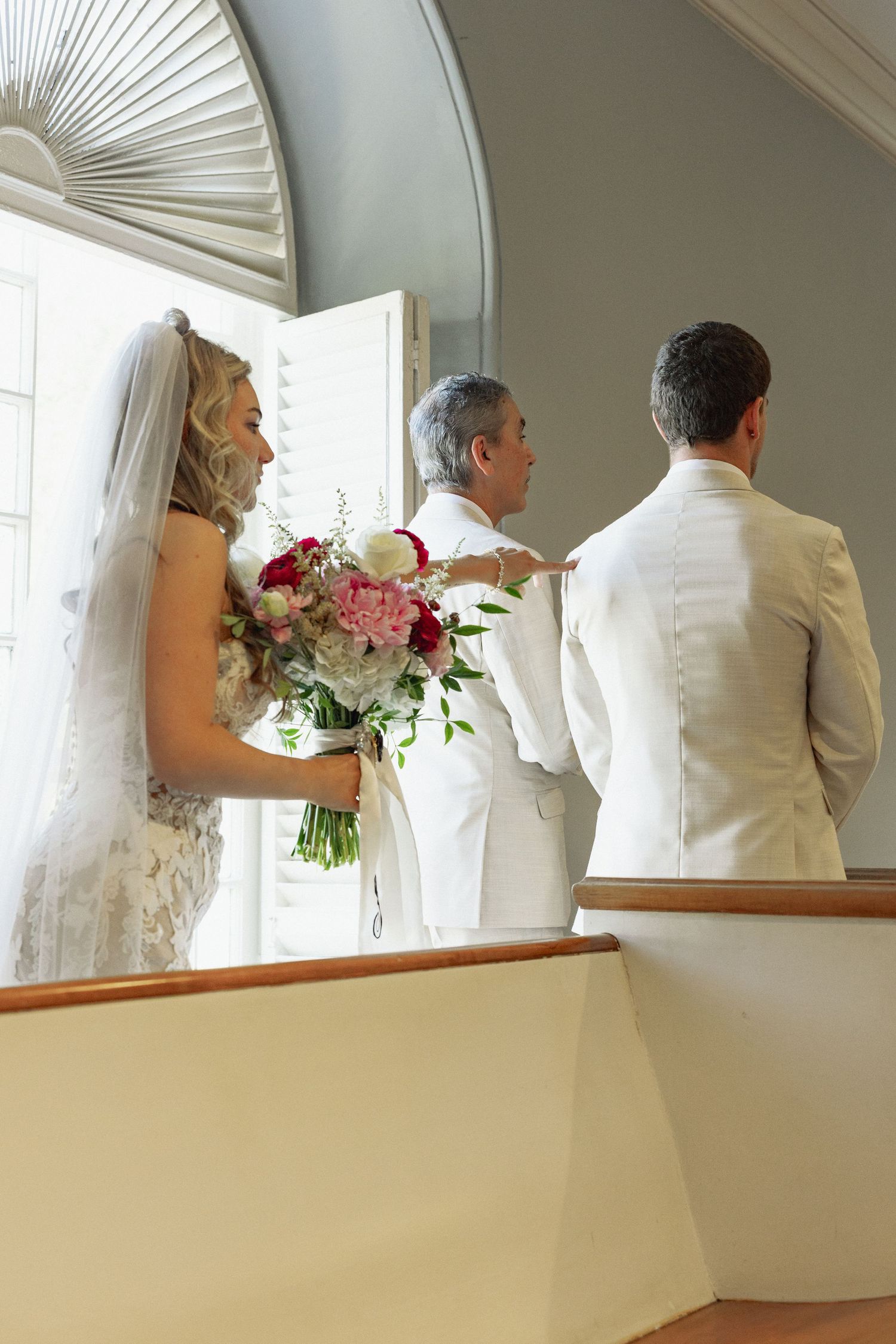 Bride holding pink bouquet stands with groom and officiant near arched window before wedding ceremony.