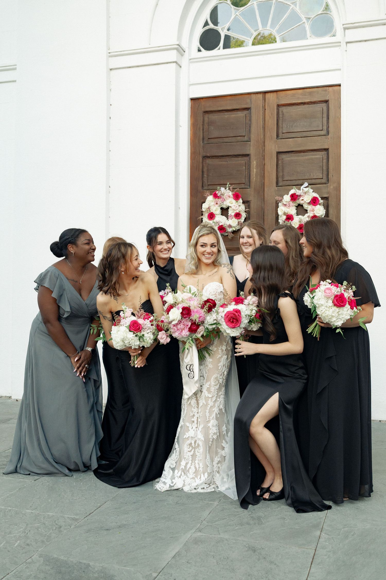 Bride in lace gown with bridesmaids in black and gray dresses holding red and white bouquets outside a white church.