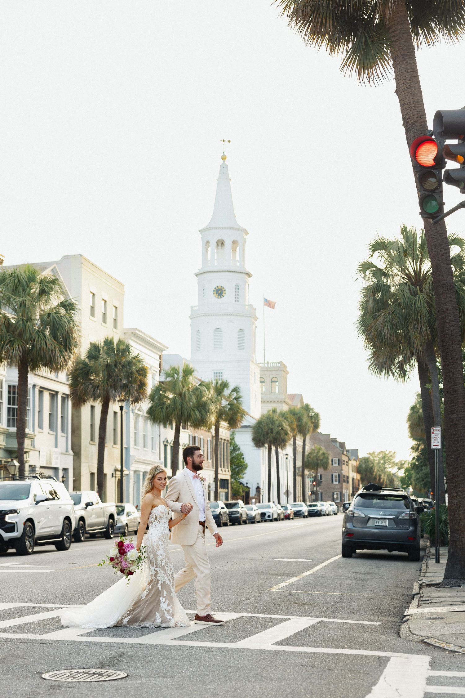 Bride and groom cross street in Charleston SC with iconic white church steeple and palm trees in background.