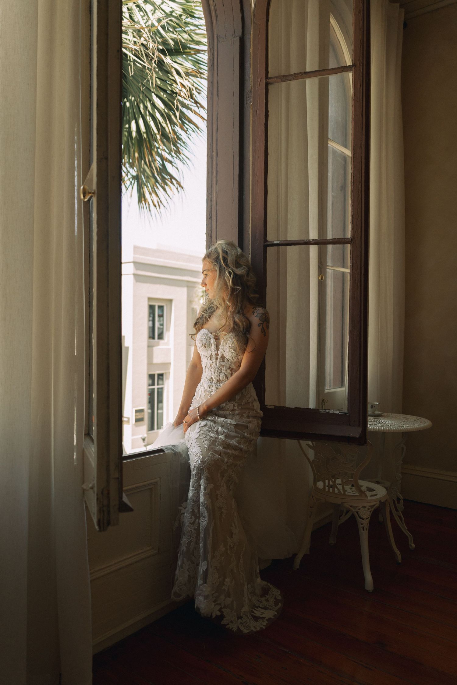 Bride in lace wedding gown sitting on windowsill gazing outside at palm trees in elegant room.