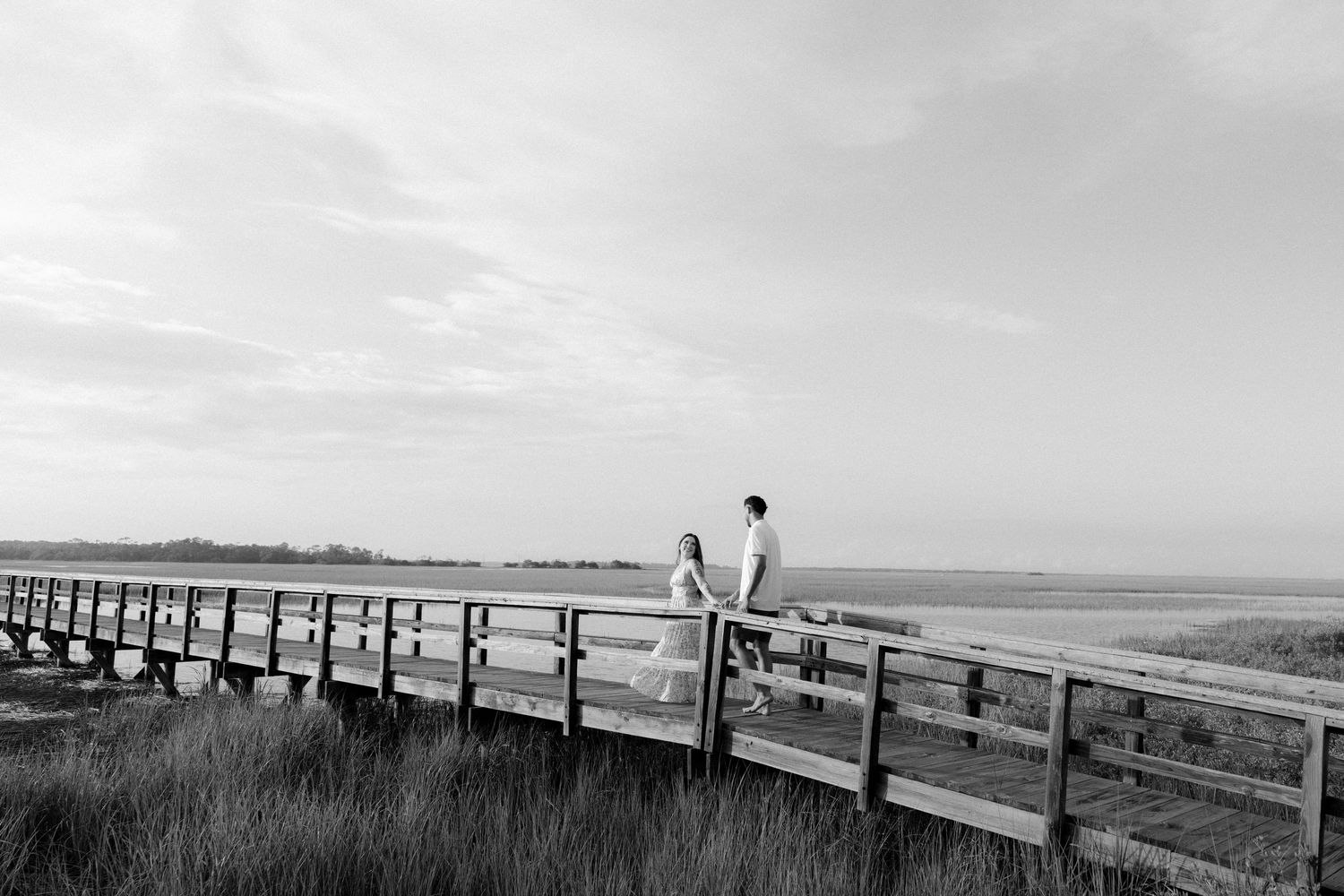 Black and white photo of a couple holding hands on a wooden boardwalk overlooking a serene coastal marsh landscape.