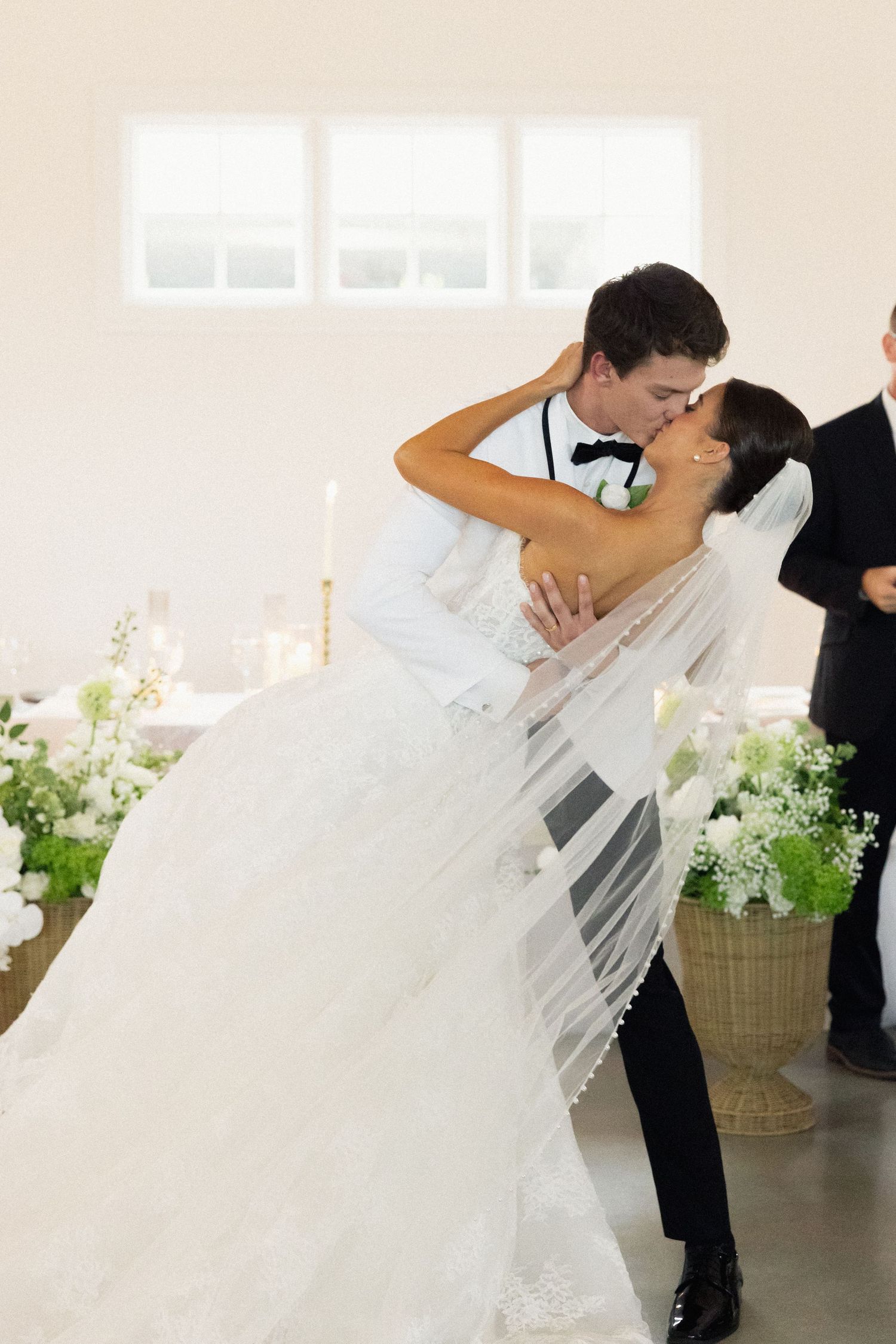 Bride and groom share romantic dip kiss at altar surrounded by white floral arrangements.