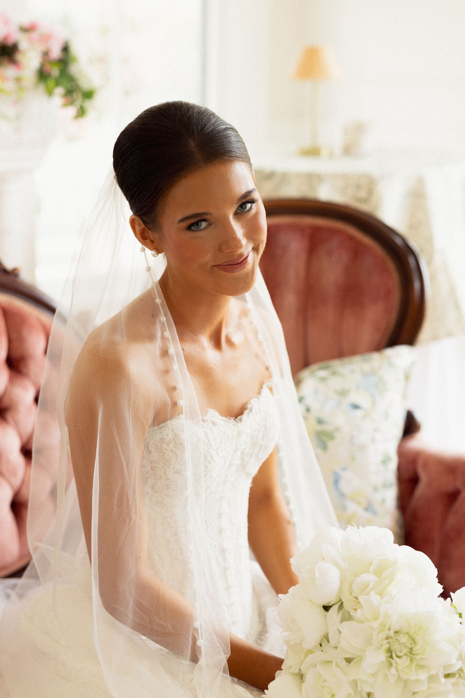 Elegant bride in a white lace gown and veil holds a bouquet of white flowers while seated in a vintage chair.