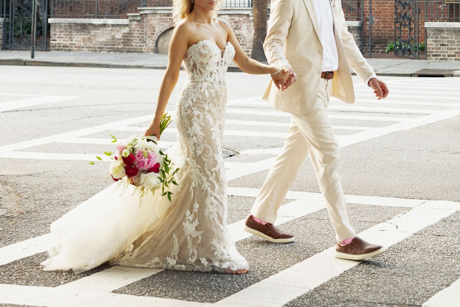 Bride in lace gown holding bouquet crosses street with groom in cream suit on wedding day.