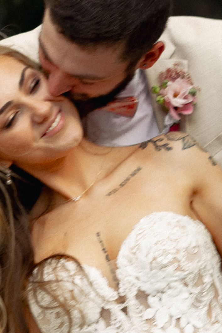 A bride in lace gown smiles as groom in suit with floral boutonniere kisses her cheek at wedding.