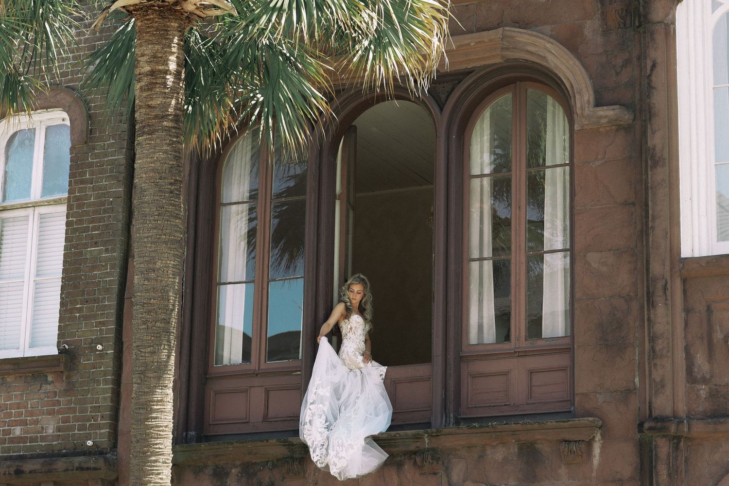 Bride in flowing white gown standing on ornate brick building ledge beside palm tree.