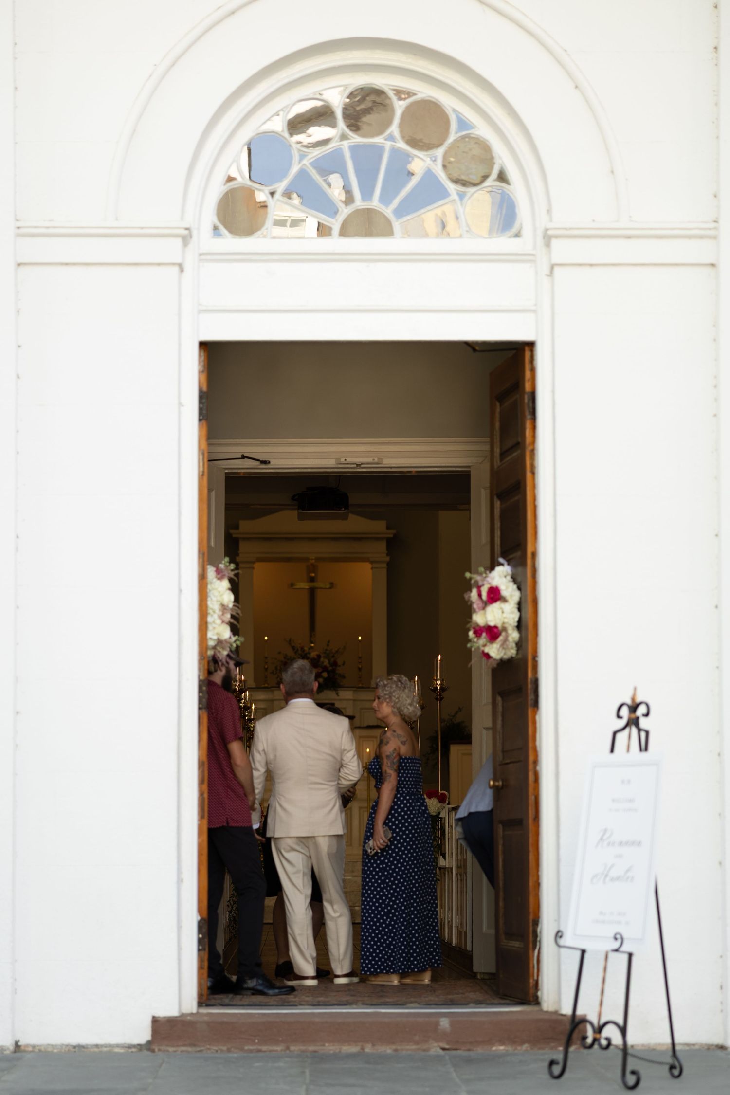 Guests arriving at a white church entrance with wooden doors, floral decorations, and a wedding welcome sign on an easel.