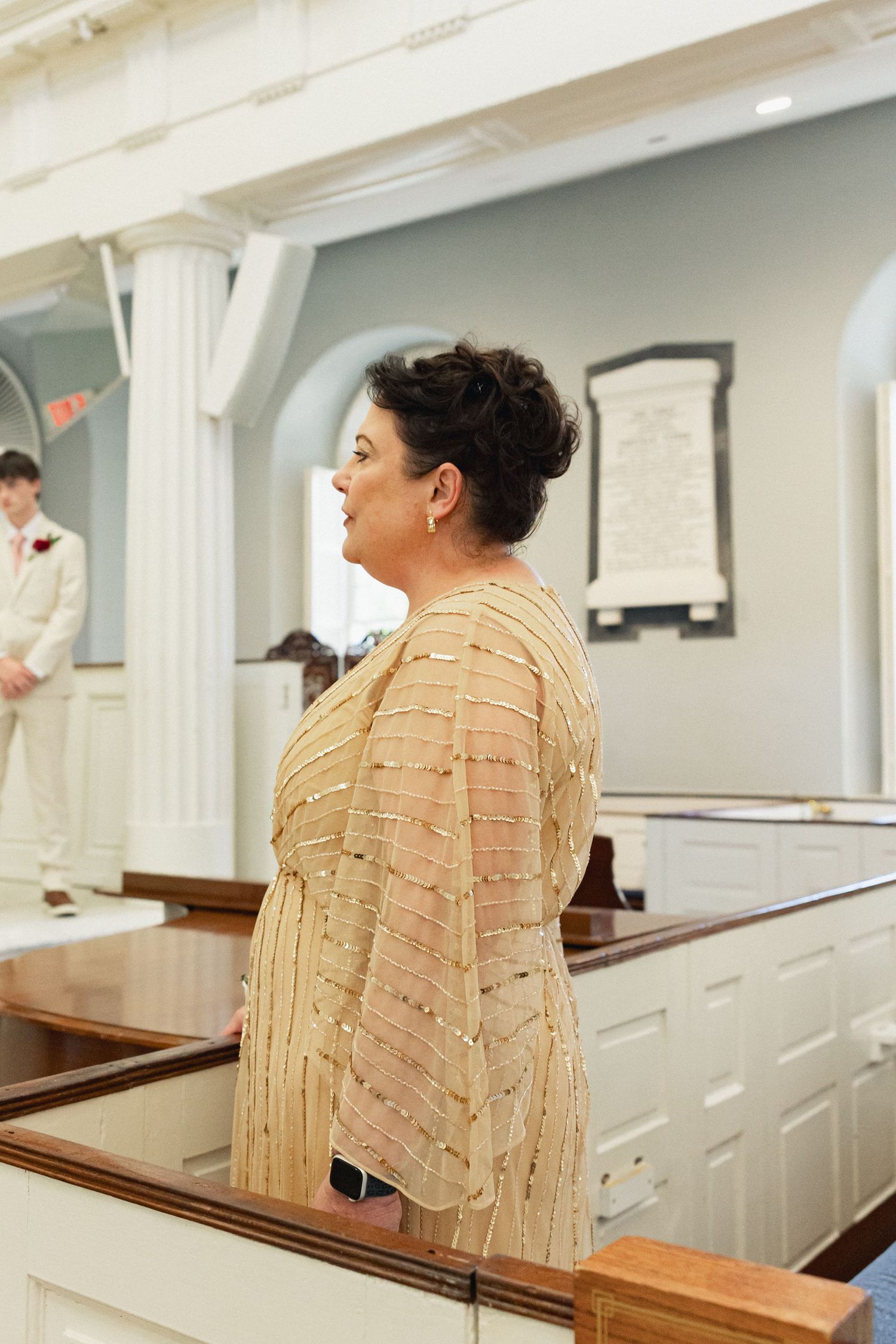 An elegant woman in a gold beaded dress stands in a church during a wedding ceremony.