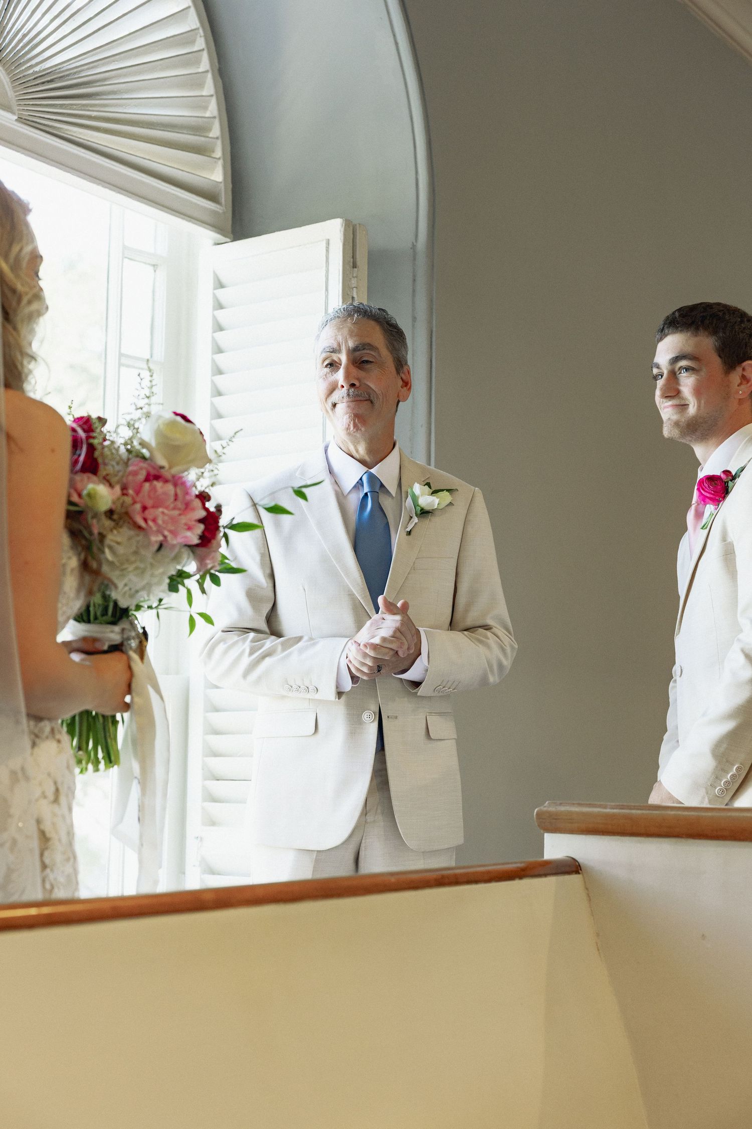 Smiling officiant in tan suit clasps hands facing bride and groom at elegant wedding ceremony.