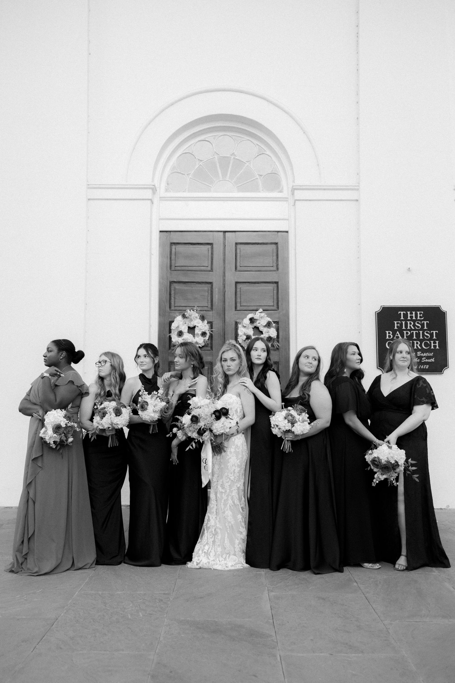 Bride and bridesmaids in black gowns pose outside First Baptist Church holding floral bouquets in black and white photo.