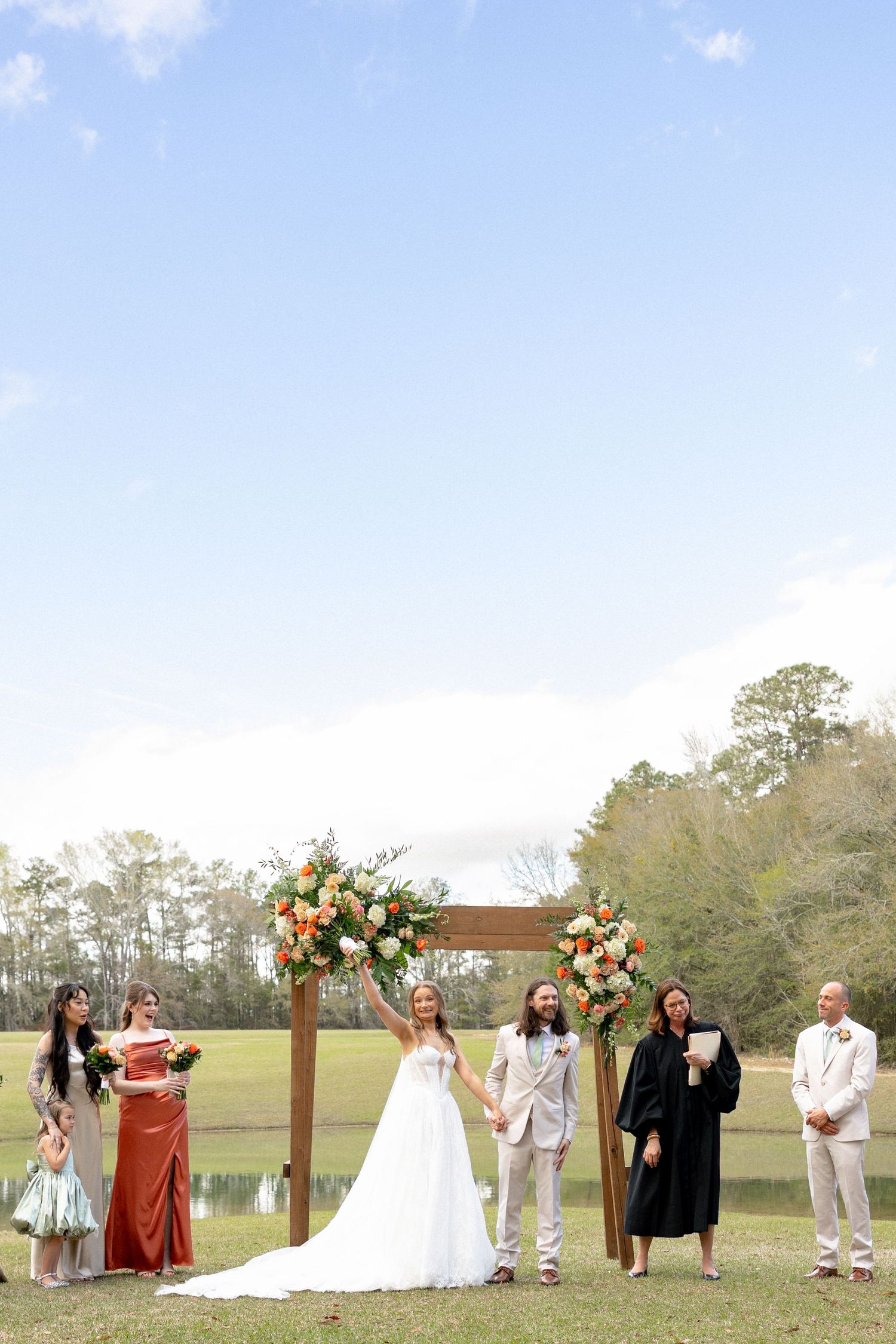 Wedding party celebrates at rustic outdoor ceremony beneath wooden arch adorned with orange and white flowers.