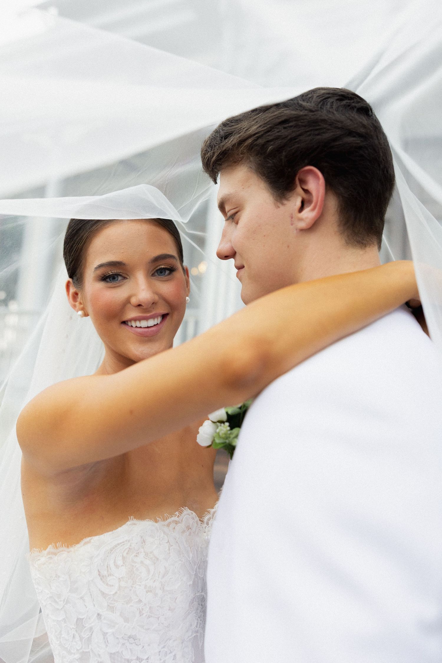 A smiling bride in a white lace gown and veil embraces her groom in a white suit during their wedding.