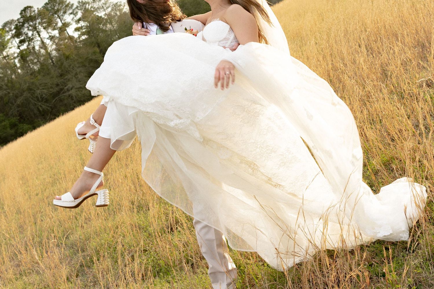 Groom carries bride in flowing white wedding dress through tall golden grass field.