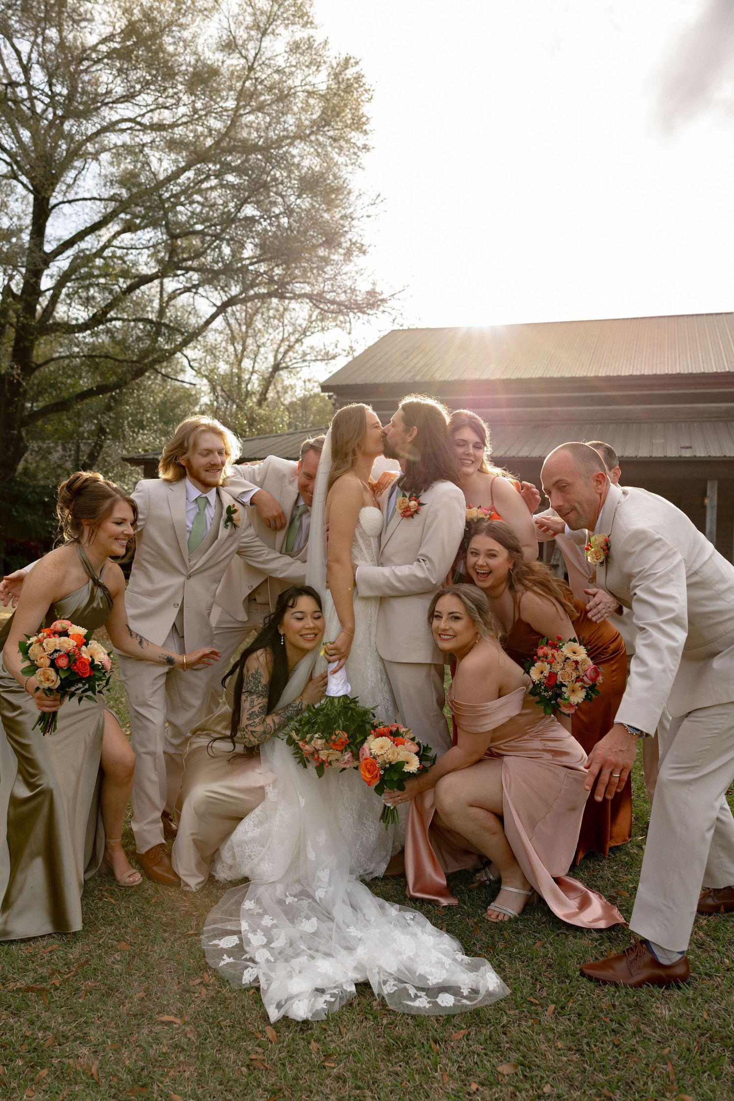 Wedding party clusters around kissing bride and groom in golden sunlight beside a rustic barn venue.