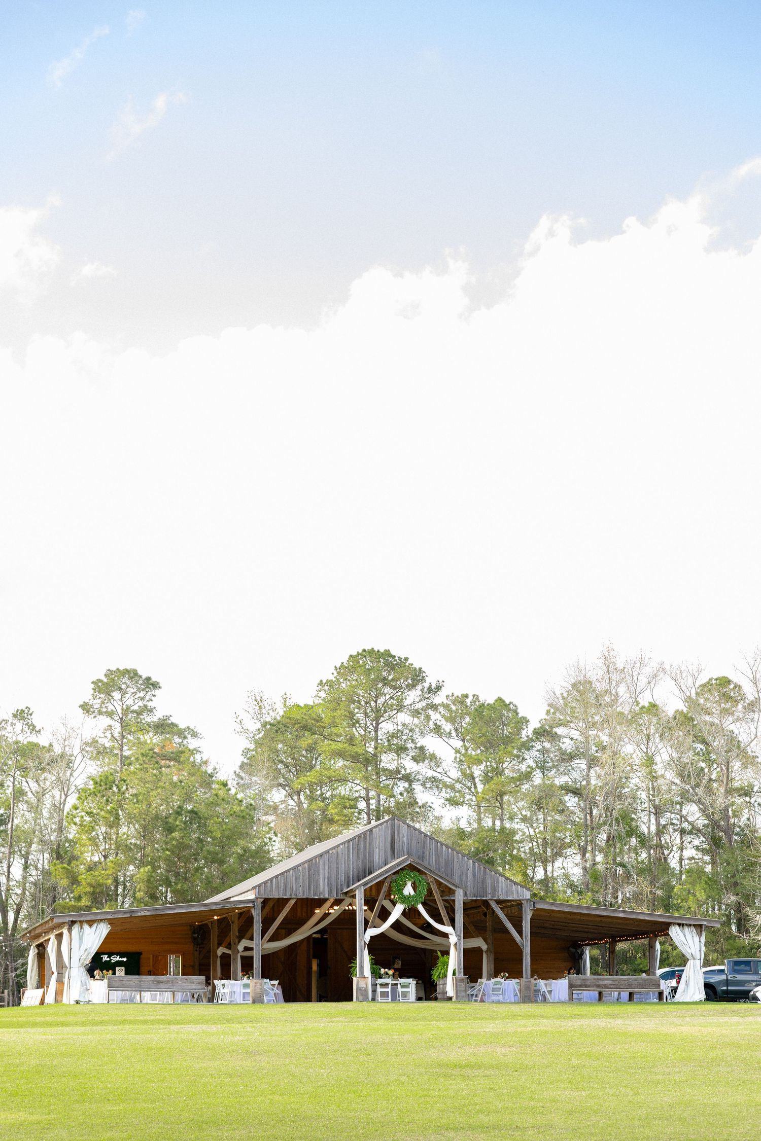 Rustic wooden barn wedding venue surrounded by tall pine trees under a bright blue sky with white clouds.