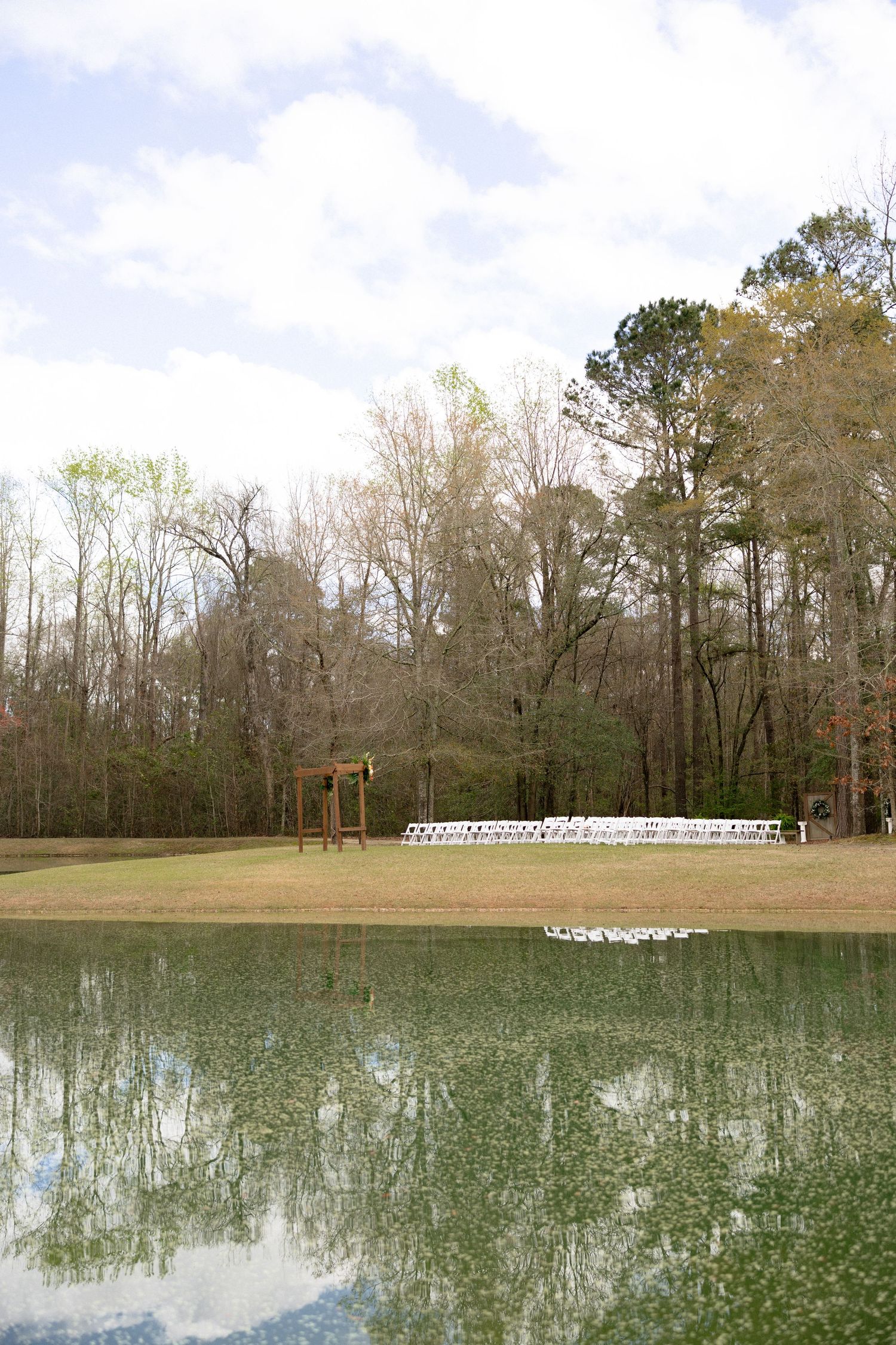 Serene outdoor wedding ceremony setup with white chairs and wooden arch reflected in a tranquil pond.