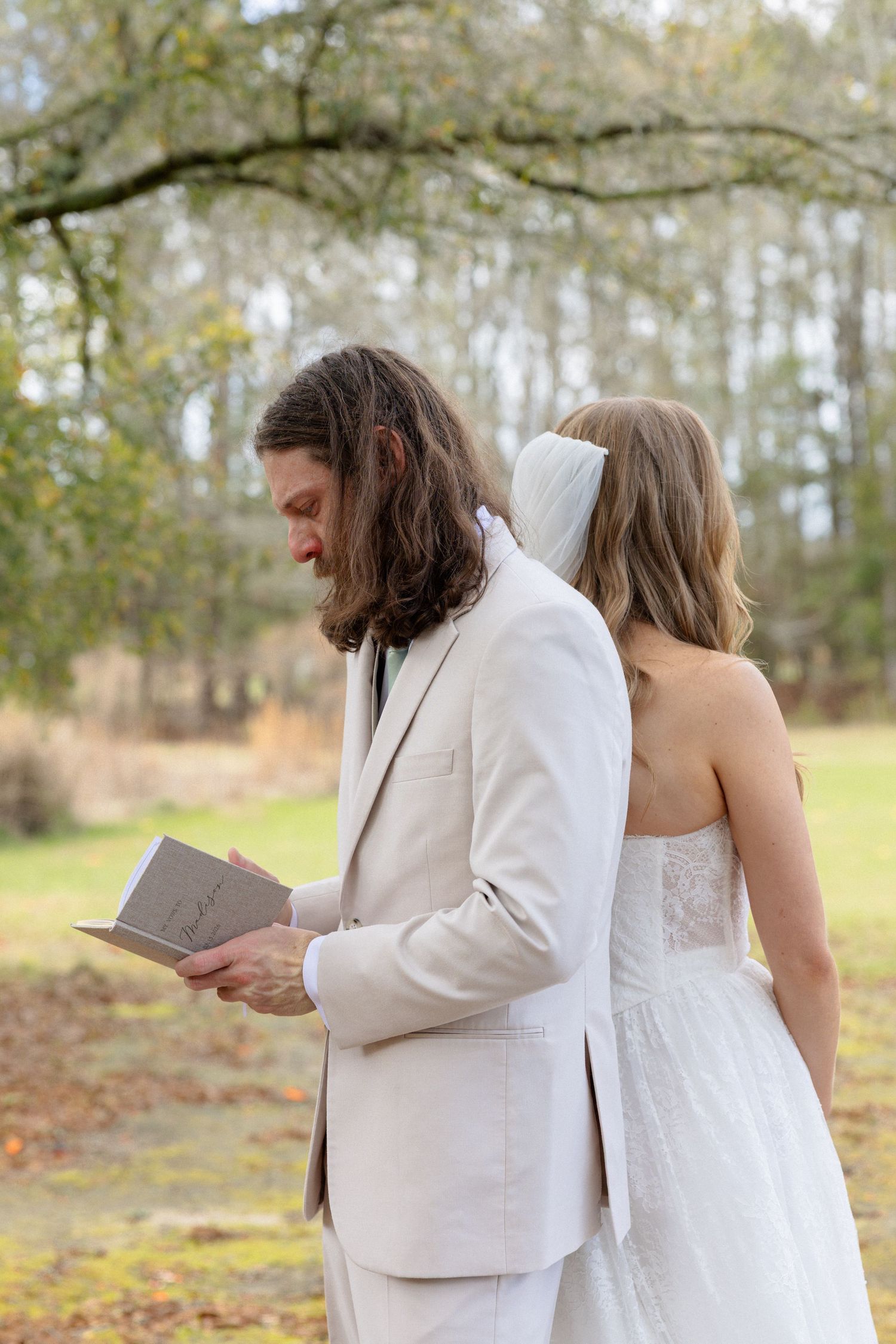 Groom in beige suit reads vow book back-to-back with bride in lace wedding dress during first look.