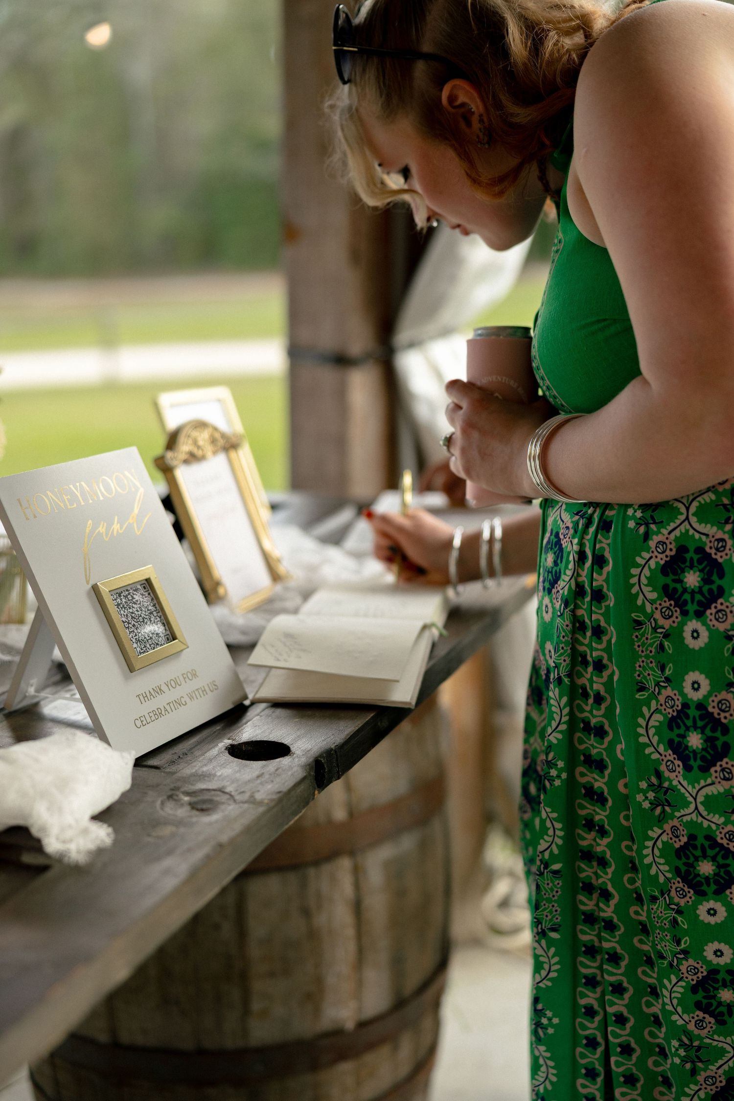Wedding guest in green floral dress signing the guestbook at a rustic barrel table.